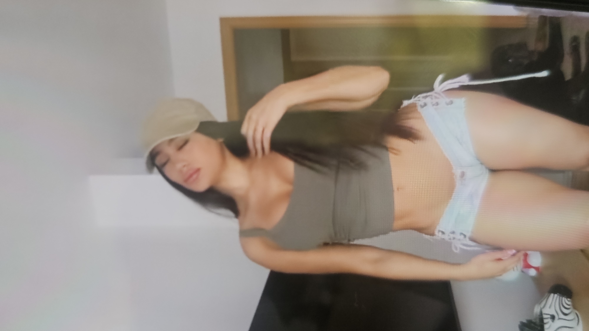 A young adult female, appearing East Asian, poses indoors within a room with light-colored walls. She wears a beige baseball cap, an olive green ribbed crop top exposing her midriff, and light-wash denim shorts featuring white lace-up detailing with bows on both sides. Her long, dark hair is styled down, and her head is tilted backward to her left with closed eyes. Her left hand rests near her left shoulder. To the left, a dark flat-screen television is partially visible, alongside a light-colored wooden table supporting a white zebra-striped object and a small red and white plush toy. In the lower right background, a dark-furred dog is partially visible, appearing to be lying down. A light-colored wooden door frame is present on the right side of the image. The location is Unknown, Unknown.