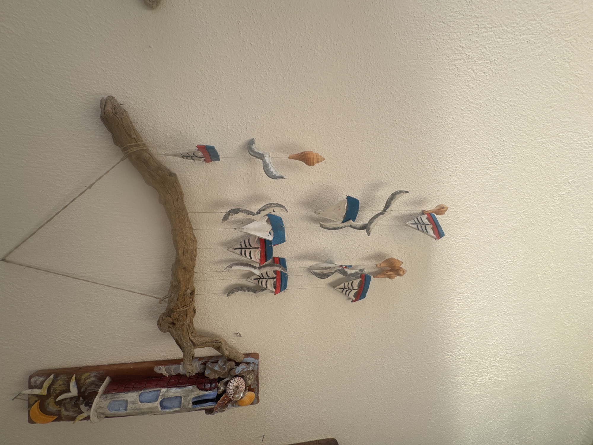 This is a medium shot, eye-level, indoor photograph of a decorative hanging piece mounted on a textured, off-white wall. The hanging piece consists of a weathered piece of driftwood suspended by a thin rope. From the driftwood, multiple strings descend, each adorned with miniature wooden sailboats, some with red and blue hulls and white sails, and others with all white sails. Interspersed with the sailboats are small, tan-colored shells and a grey seagull figurine. To the left of the driftwood is a vertical, wooden decorative plaque depicting a lighthouse with blue windows, a red brick pattern, a crescent moon, and several white birds in flight, all rendered in a folk art style. Small shells are also attached to the bottom of this plaque. The overall scene suggests a coastal or nautical theme. The lighting appears to be ambient, coming from an unseen source. The photograph was taken in Ammouliani, Greece.