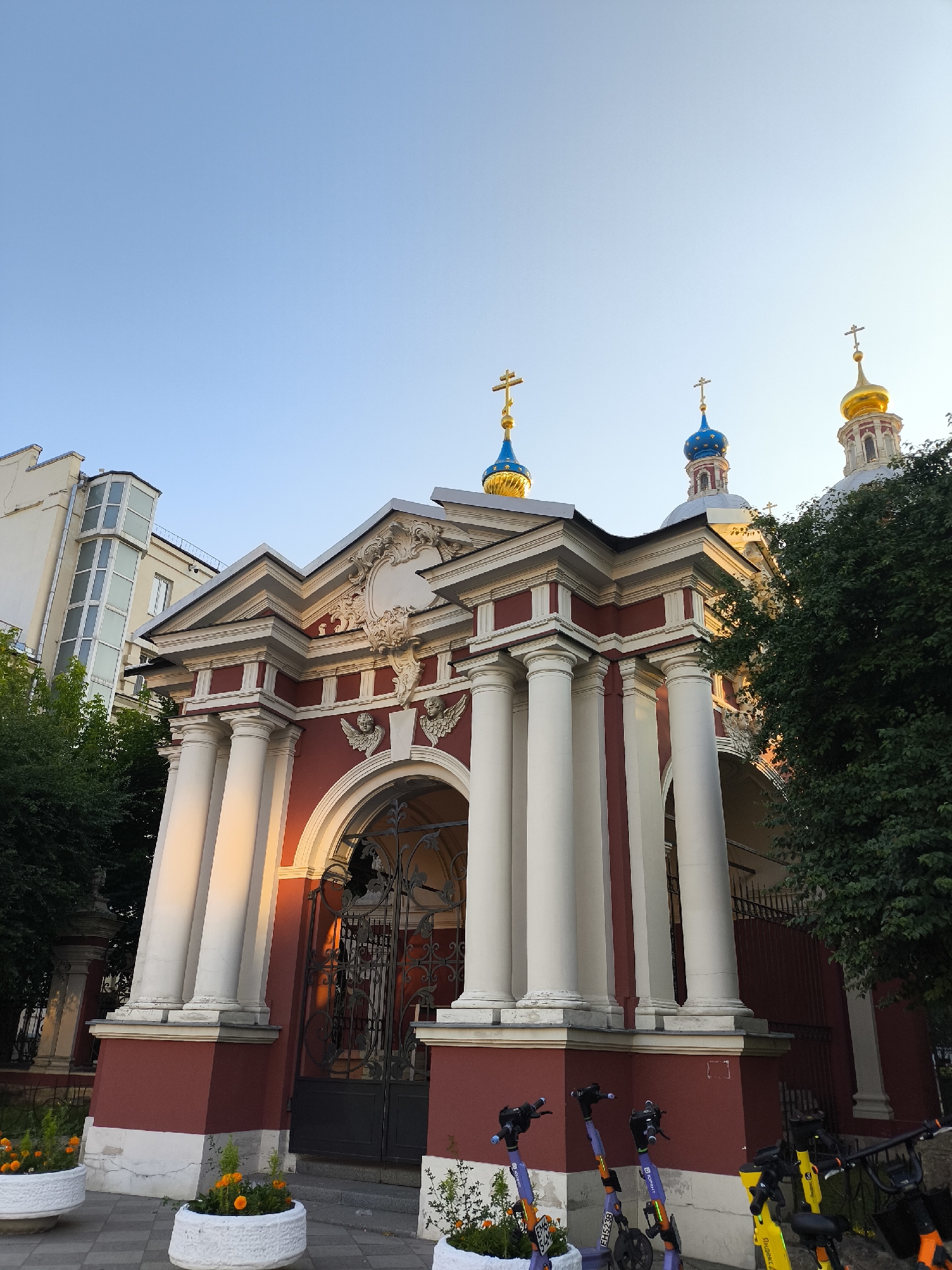 This image captures the exterior of a richly decorated building, likely a church or a historical monument, bathed in the warm glow of what appears to be late afternoon or early morning sun. The building features a striking facade in deep red and white, adorned with ornate columns, elaborate carvings, and classical architectural elements like pediments and cherubs. Three onion domes with crosses on top are visible against a clear, pale blue sky, suggesting fair weather.
In the foreground, several parked electric scooters are lined up, adding a modern contrast to the historic architecture. Two of the scooters are purple, and at least one is yellow. White planters filled with orange flowers are placed along the sidewalk, introducing touches of vibrant color and nature. To the left of the main building, a modern apartment building with a distinctive glass-enclosed section rises into the frame, indicating an urban setting where historical and contemporary structures coexist.
The sunlight highlights the architectural details, creating strong contrasts and shadows, and casting a warm hue on the red walls and the white columns. The overall scene evokes a sense of tranquility and historical grandeur, juxtaposed with the everyday presence of modern transportation. No explicit text is legible in the image, apart from some branding on the scooters, which are too small to decipher.