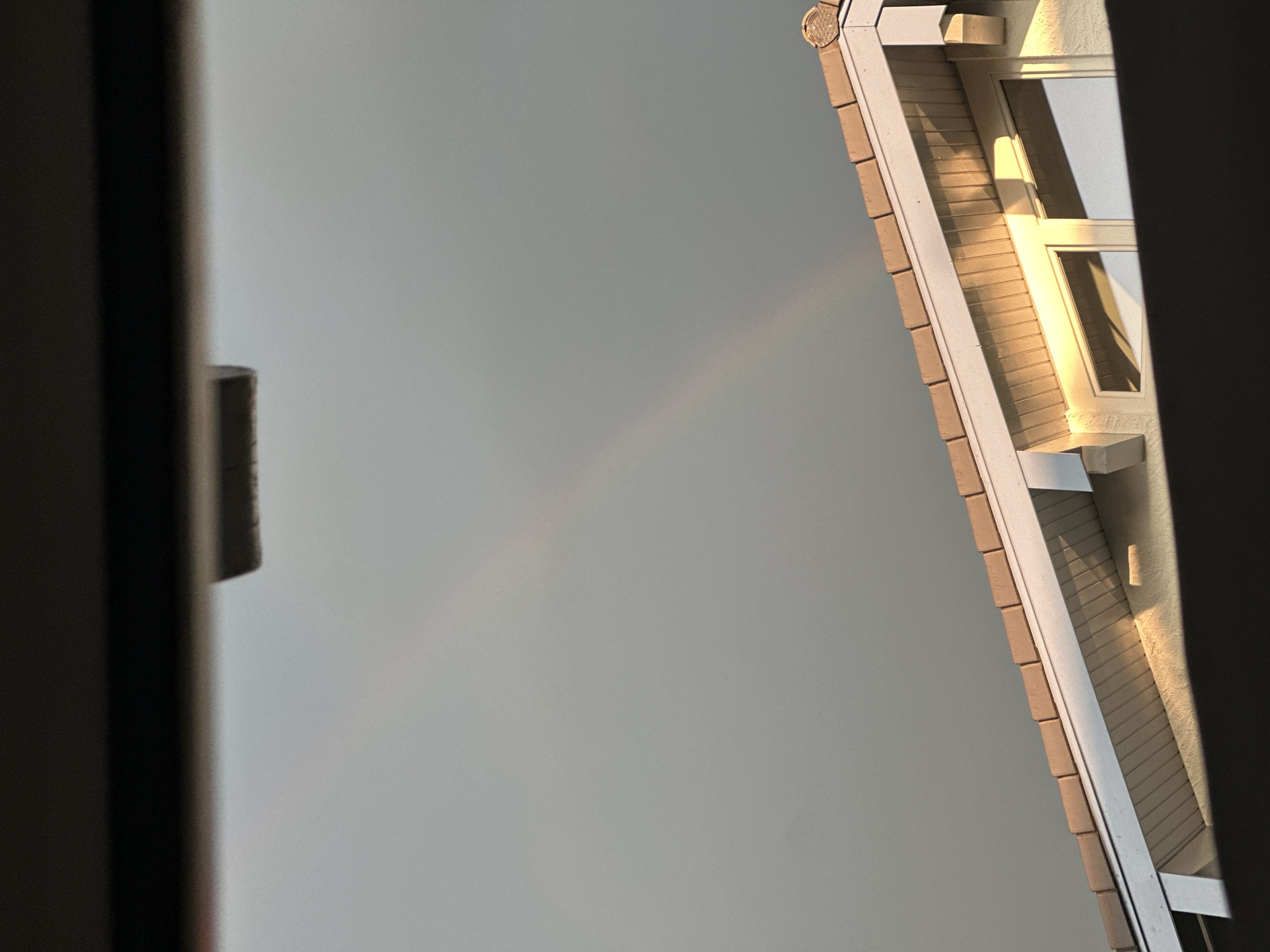 The image shows a partially obscured view of a house roof with tan tiles against a grey sky. A faint rainbow arc is visible in the upper portion of the sky. The lower part of the image is filled with a dark, blurred foreground that obscures the view of the house and sky. There are no people or animals visible in the image. The location in the city of Bad Endorf, Germany is not discernible from the image.