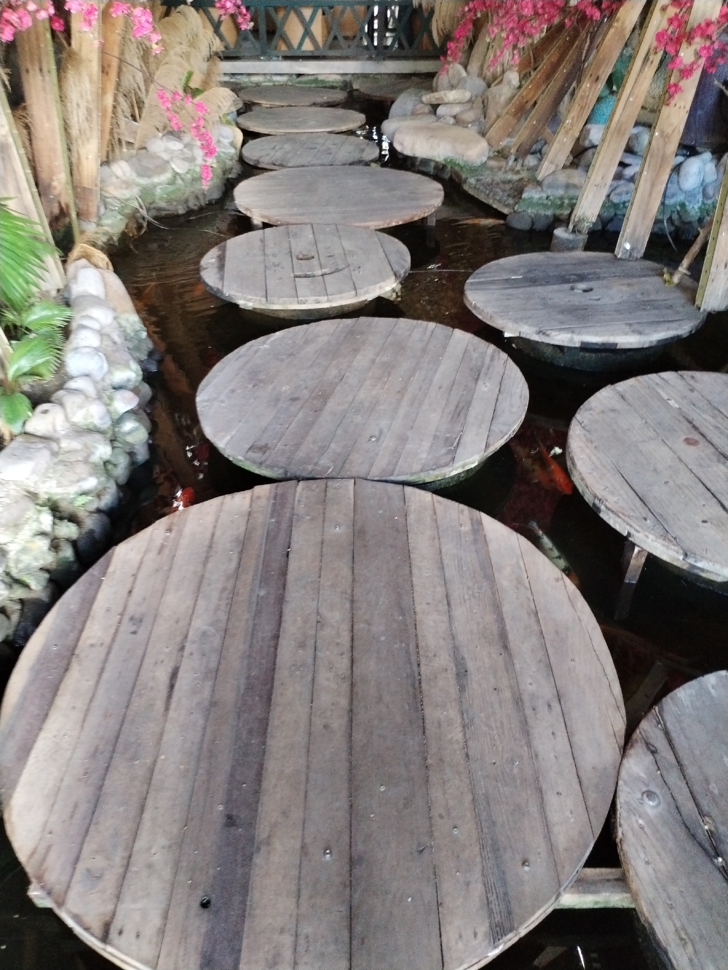 The image depicts a series of large, circular wooden platforms placed in a pond or shallow body of water. These platforms serve as stepping stones, creating a pathway across the water. The platforms are constructed from wooden planks, showing signs of wear and aging. Surrounding the pond are decorative elements including artificial plants, rocks, and wooden supports. The water is clear enough to reveal koi fish swimming beneath the platforms. The location is identified as Bac Nha Trang Ward, Vietnam.
