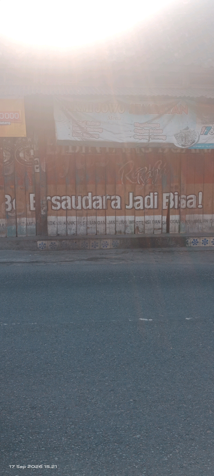 This is a low-angle, slightly hazy, outdoor photograph of a street scene in Wedi, Indonesia. The primary subject is a rustic-looking wooden wall that serves as a backdrop for signage and graffiti. The wall is constructed from vertical wooden planks of varying widths and shades of brown, some of which are painted over with a mural.
At the top of the wall, a banner with Indonesian text is visible, though it is largely obscured by a bright, overexposed sky or light source. The text appears to be the name of a business, possibly a restaurant, with "NAKMI JOWO KEMBATAN" being partially decipherable. Below this banner, a menu list is partially visible, with items like "Bakmi Rebus," "Bakmi Goreng," and "Nasi Goreng."
Dominating the lower portion of the wall is a large, white slogan in a bold, blocky font that reads "Bersaudara Jadi Bisa!". This translates to "Brothers/Siblings Become Capable." Below this slogan, another line of text is present, but it is difficult to read due to its size and the texture of the wall. Snippets like "ABKAN KANKER, SERANGAN JANTUNG IMPOTENSI DAN GANGGU KEHAMILAN DA" can be discerned, suggesting it might be an advertisement for traditional medicine or remedies.
To the left of the main slogan, a vertical sign with a yellow background and black text reads "0.000 Batang," potentially indicating a price or quantity. Faintly visible on the left side of the wall are logos of what appear to be beverage brands like "BINTANG."
The wall is adorned with graffiti, including a stylized red script that is difficult to identify. The bottom edge of the wall transitions into a concrete curb with decorative blue and white tile patterns. The foreground of the image is dominated by a dark grey asphalt road, which appears to be slightly wet or reflective. The timestamp "17 Sep 2026 15.21" is visible in the bottom left corner, indicating the date and time the photograph was taken. The overall atmosphere is somewhat gritty and urban, with the bright light source creating a diffused and slightly ethereal quality.