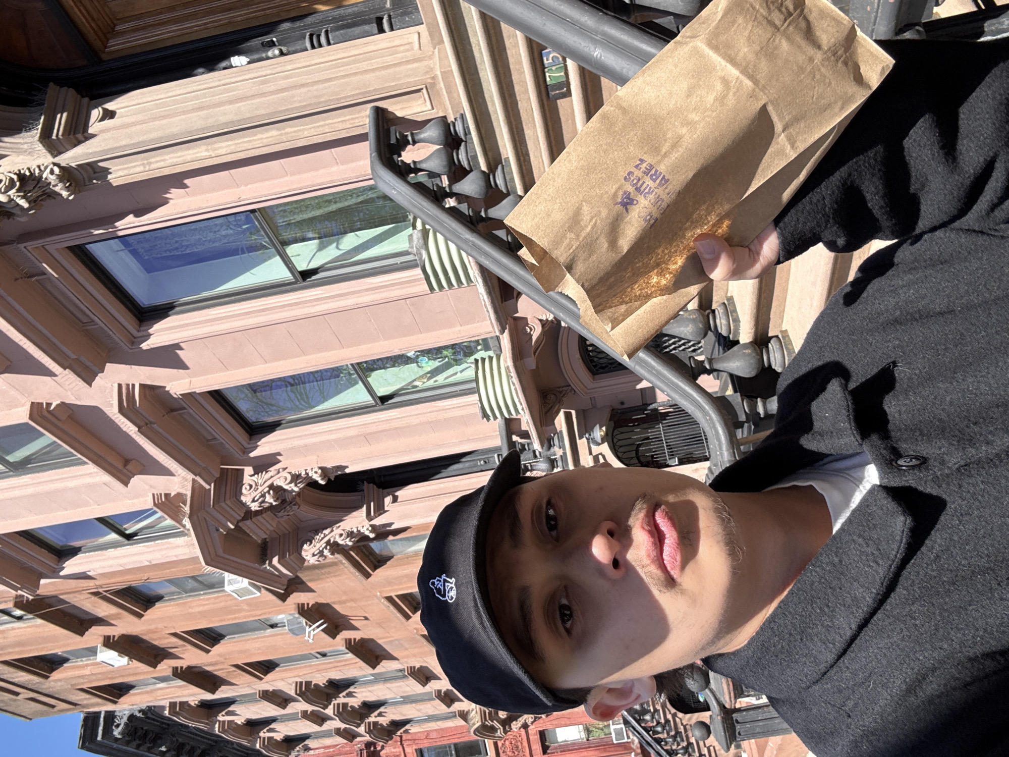 The image shows a person standing on a sidewalk in front of a brick building in New York City. The person is holding a brown paper bag with writing on it. The building behind the person has windows and decorative architectural elements. The location is identifiable as New York City due to the architectural style of the building, which is common in many neighborhoods.