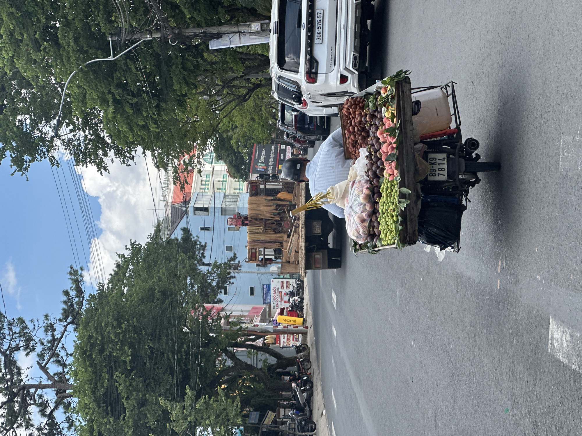 The image depicts a street scene in Lâm Viên - Đà Lạt, Vietnam, on a cloudy day. In the foreground, a person wearing a light blue and white striped shirt and a dark helmet is riding a motorcycle. The motorcycle is outfitted with a wooden cart loaded with a variety of fruits. Visible fruits include green round fruits, red-skinned fruits, and darker purple fruits, possibly mangosteens. A large white plastic bag filled with more produce is also on the cart.
Behind the motorcycle, a flatbed truck is stopped, carrying stacked bundles of what appears to be fibrous material, possibly dried plants or wood. Further down the road, several cars are in traffic, including a white Ford Everest SUV with the license plate 30K-576.57. The surrounding environment includes buildings with colorful facades, primarily blue and pink, with red roofs. Lush green trees line the street, and overhead power lines are visible against the sky. Motorbikes are parked on the side of the road in the middle ground. The road surface is asphalt, with white lane markings.