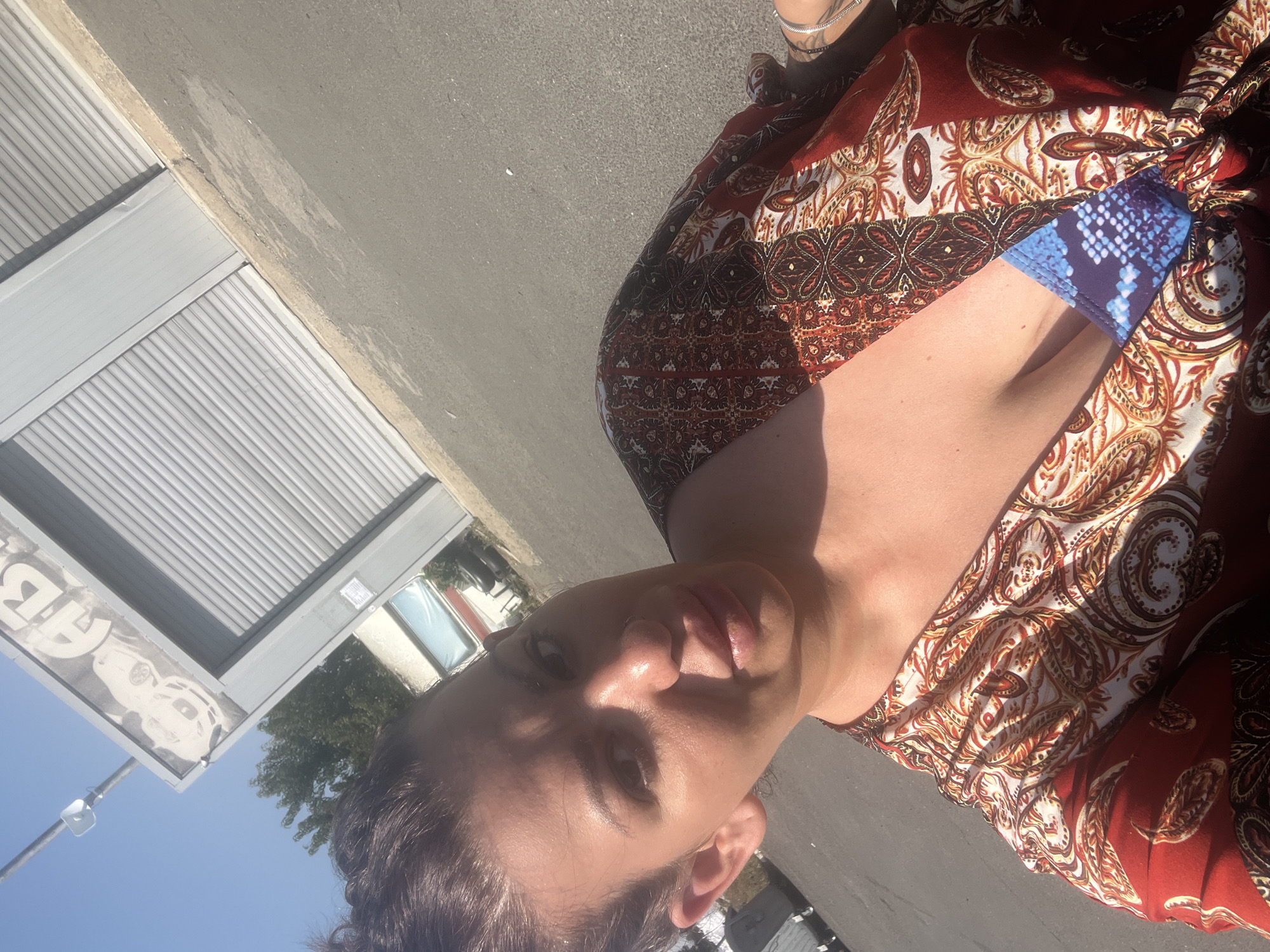 A woman is taking a selfie outdoors, in a city. She is wearing a red, orange, and white patterned dress over a blue and white patterned swimsuit. The background shows a gray roller door on a building and an asphalt ground. The sky is clear and blue. The image appears to have been taken during daylight.