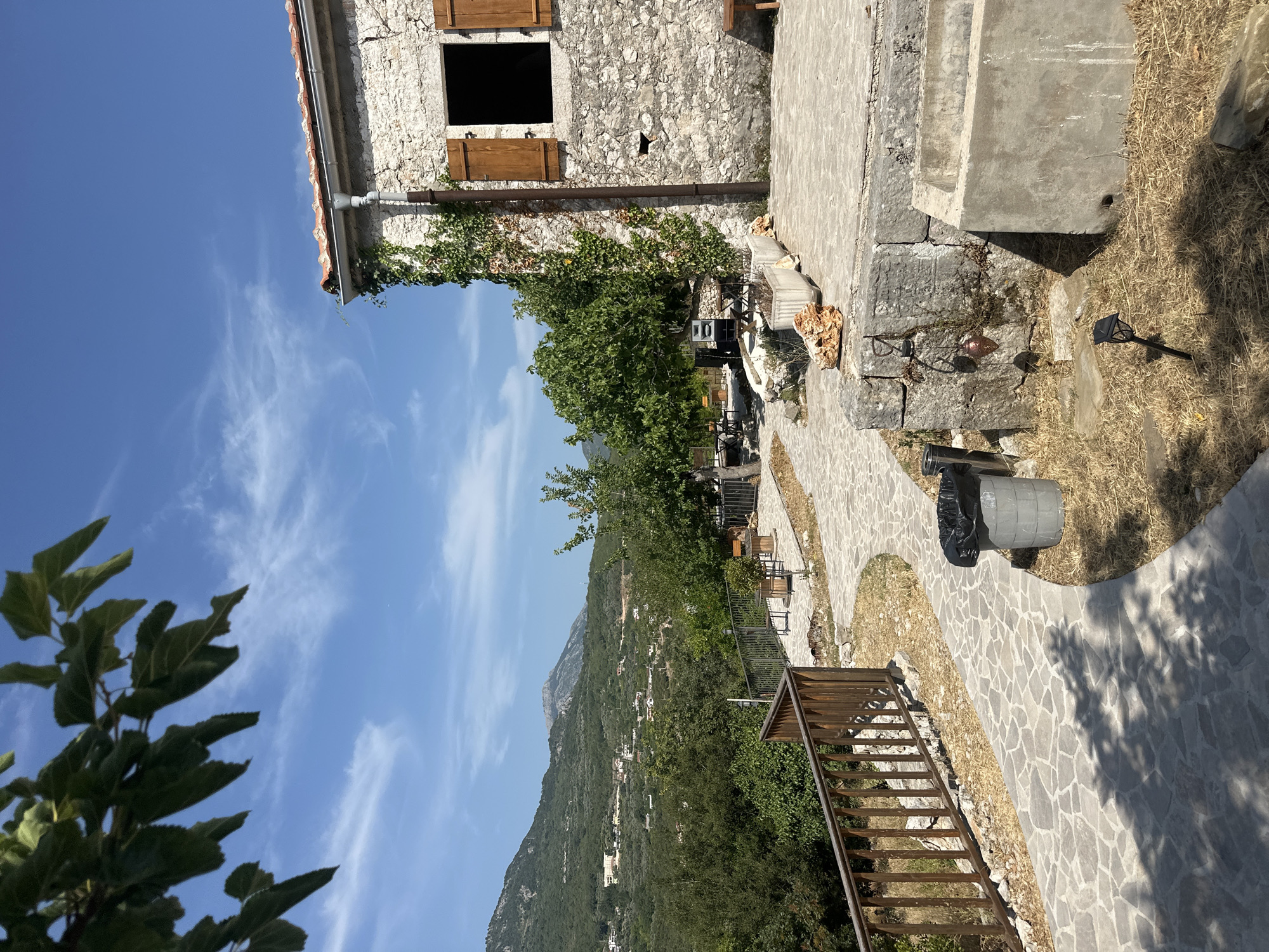 The image depicts a scenic view of a stone building and its surroundings in Pečurice, Montenegro. In the foreground, a stone pathway winds through dry grass and leads towards a rustic stone building. The building has wooden shutters on a large window and is partially covered in ivy. Adjacent to the building is a concrete structure, possibly a water trough. A metal trash can with a black bag is positioned near the pathway. In the mid-ground, a metal fence runs along the edge of a patio area with tables and chairs, overlooking a lush green landscape of trees and rolling hills. In the background, a mountain range stretches across the horizon under a bright blue sky with scattered white clouds. The overall scene conveys a sense of rural tranquility and natural beauty.