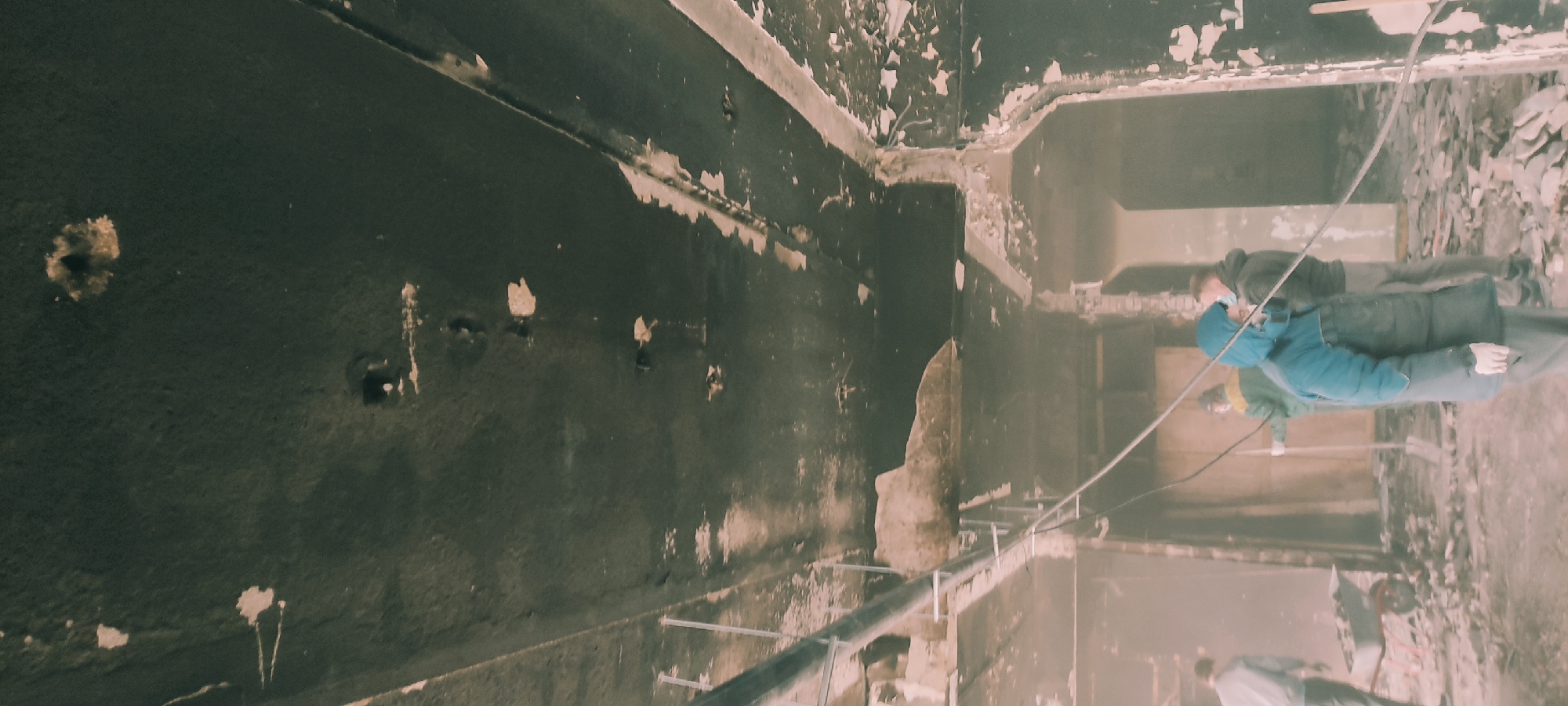 This image captures an interior scene, likely a large room or hall, in a state of significant disrepair or under demolition, located in Fryazino, Russia. The dominant feature is the dark, possibly soot-covered or heavily damaged walls, with large sections of paint or plaster peeling away, revealing lighter material underneath. The texture of the walls suggests either fire damage, severe neglect, or a process of removal.
Several individuals, appearing to be workers, are present in the right half of the frame. They are dressed in warm winter clothing, including thick jackets, hoods, and gloves, indicating a cool or unheated environment. One person in a prominent blue jacket and white gloves is visible, alongside another in a dark jacket behind them, and a third in a lighter, possibly green, top further back. They appear to be standing amidst a significant amount of rubble and debris on the floor, characteristic of a construction or demolition site.
Stretched across the mid-ground of the image are several wires or cables, which might be electrical lines or equipment used for the ongoing work. A partial view of a wheelbarrow can be seen in the lower right corner, blending with the debris. The overall lighting is dim and diffused, contributing to the somber atmosphere of the damaged space. No specific text is visible in the image. The scene strongly suggests a post-incident cleanup, renovation, or demolition project.