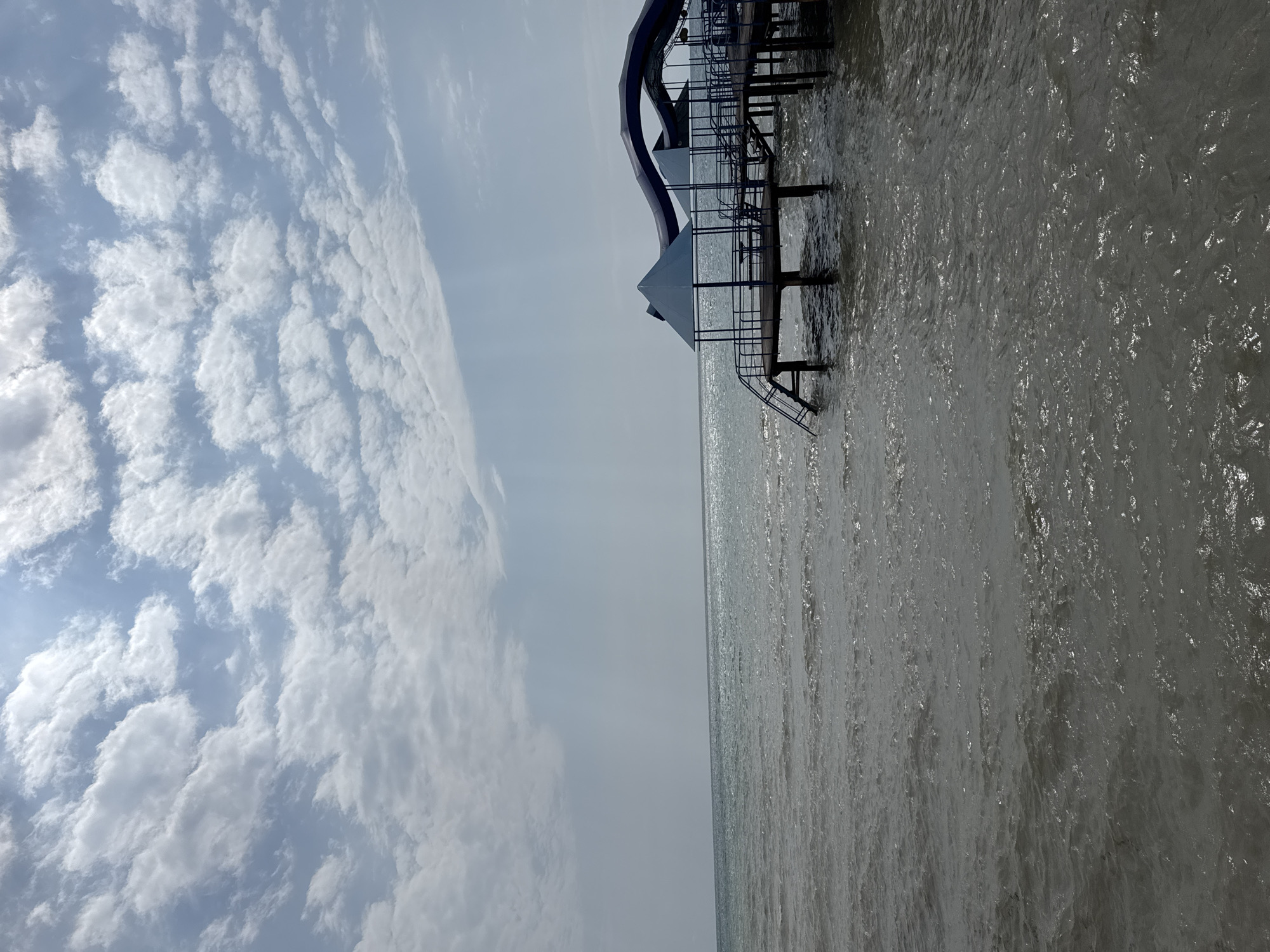 The image depicts a pier extending over a body of water, likely a lake or sea, under a partly cloudy sky. The pier is elevated on stilts and features a covered seating area with blue triangular canopies. A staircase leads down from the pier into the water. The water is choppy with visible ripples and reflects the light from the sky. The sky is a pale blue with scattered white clouds, some of which are illuminated by sunlight. The overall scene is calm and appears to be a recreational area. The location is identified as Sary-Oy, Kyrgyzstan.