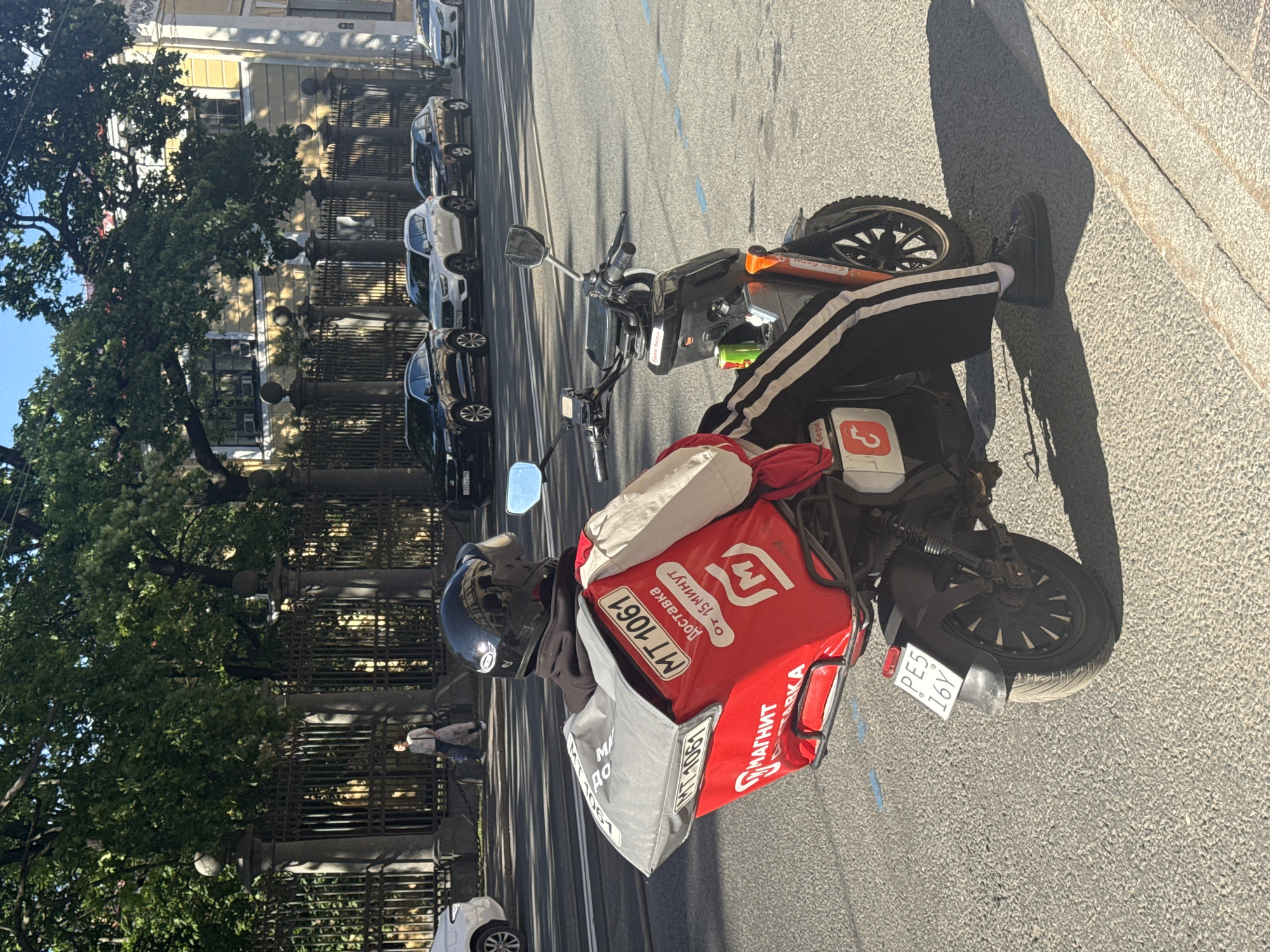A male delivery person wearing a black helmet, black hoodie, and black pants with white stripes is seated on a red and black electric scooter on a city street. He is wearing a red delivery bag with "М.КОНТАКТ" and "МАГНИТ" printed on it, as well as "Доставка от 15 минут". The scooter has a license plate that reads "PE5 16Y". In the background, there are cars parked along the street and a building with yellow walls and red roofs. Trees line the sidewalk, and a black metal fence separates the sidewalk from the street. The scene appears to be in Saint Petersburg, Russia, based on the Cyrillic lettering on the delivery bag and license plate format. The image is taken from a rear-facing perspective, focusing on the delivery person and the scooter.