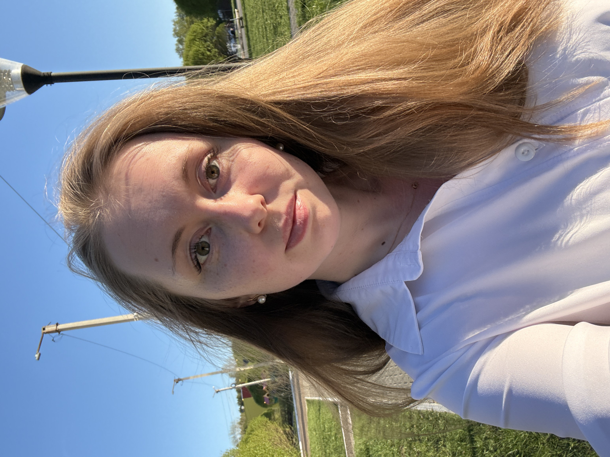 A young woman with light brown hair and fair skin is facing the camera. She is wearing a white collared shirt. The background shows an outdoor setting with a green lawn, trees, and a clear blue sky. Power lines and a lamppost are visible in the background. The location appears to be in a park or grassy area.