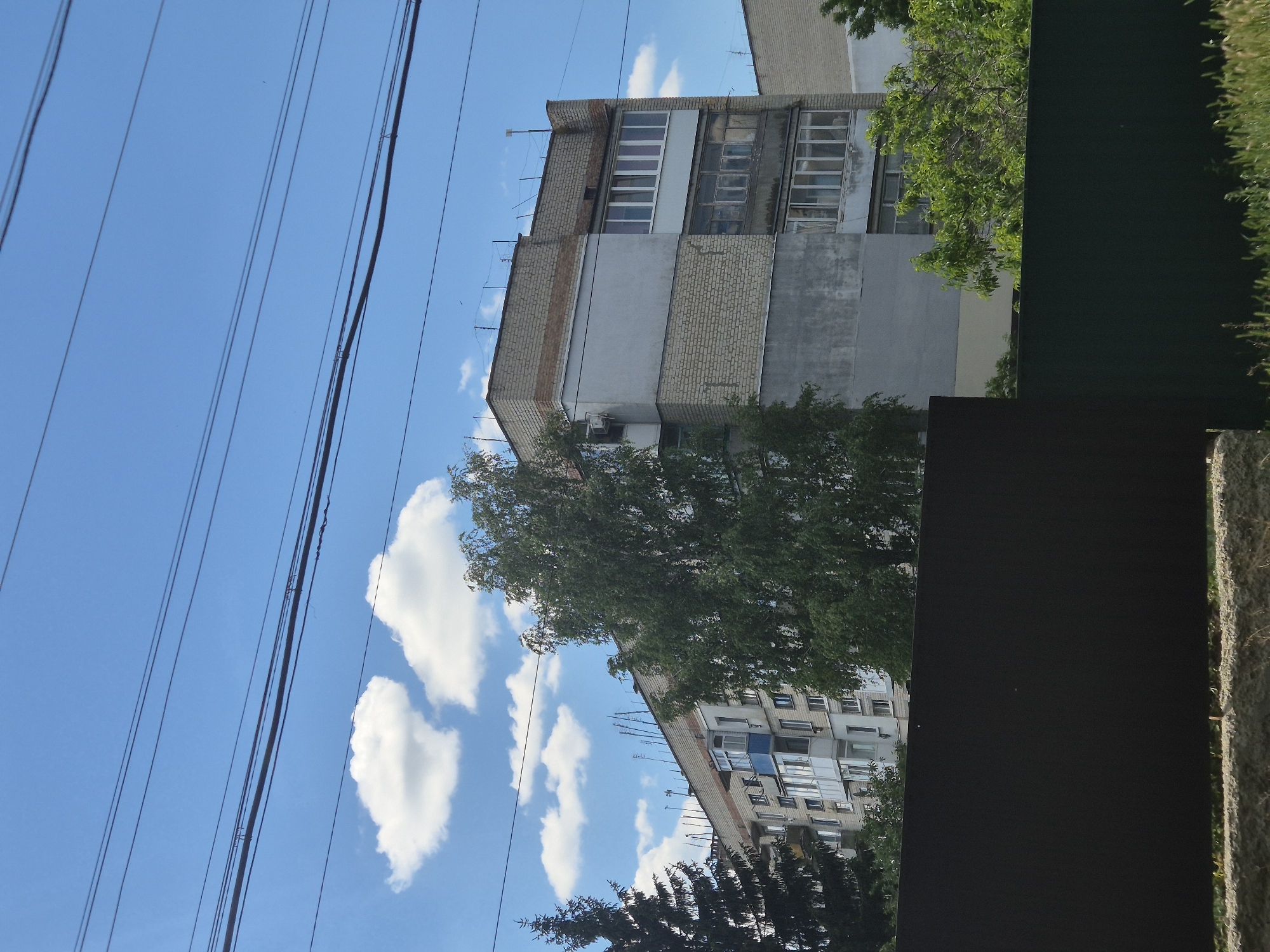 This is a medium shot, eye-level, outdoor, daytime photograph taken in Sozonivka, Ukraine. The scene depicts a multi-story apartment building in the background, constructed with a combination of light-colored brick and gray plaster. The building has numerous windows and balconies. In front of the building, there are trees with green foliage. The foreground features a dark brown fence. Overhead, multiple power lines are visible against a bright blue sky with scattered white clouds. The overall impression is of an ordinary urban residential environment on a clear day.