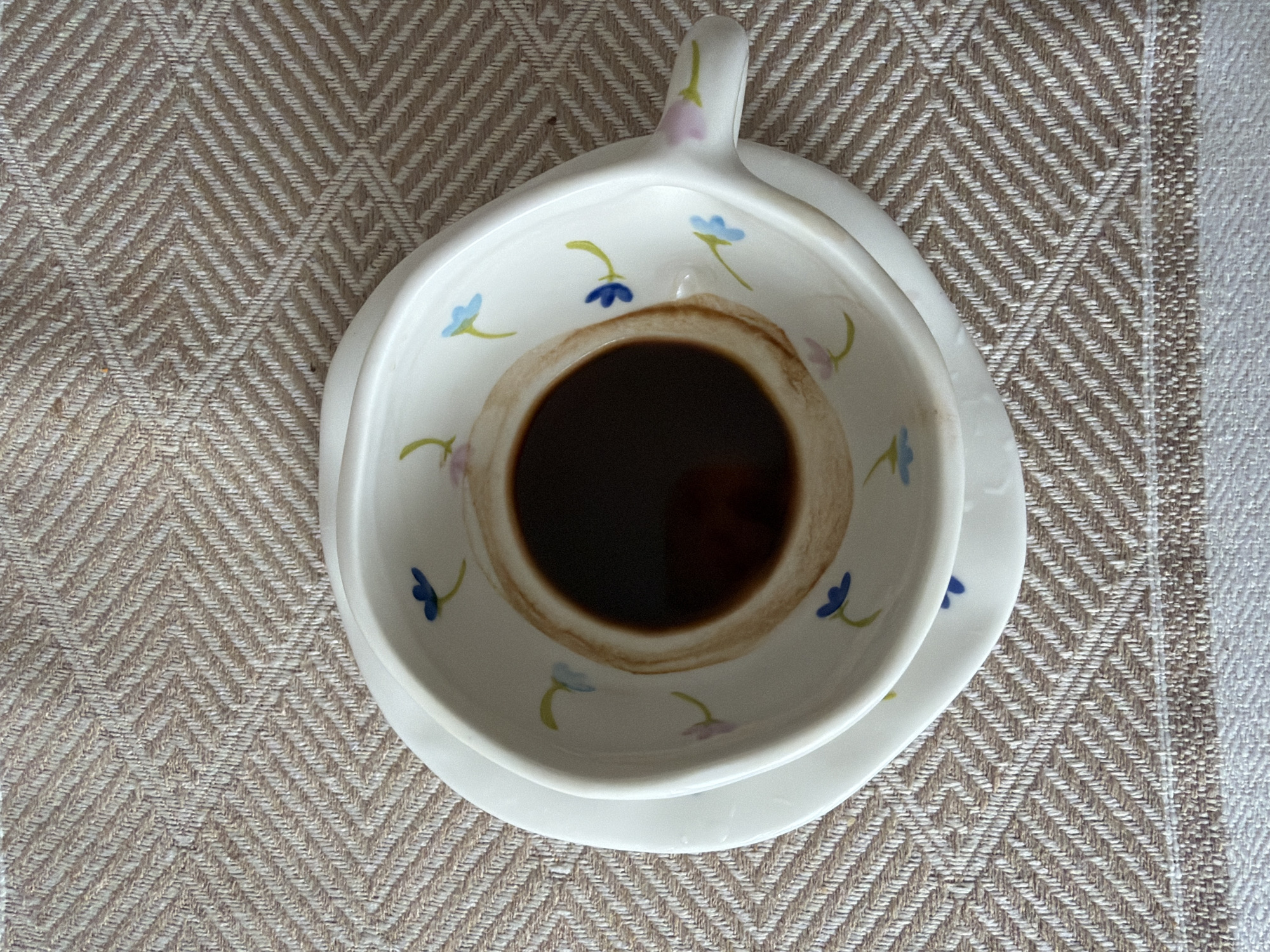 A top-down, high-angle close-up shot shows a white ceramic teacup and saucer with a flower pattern, containing a dark brown liquid, possibly coffee or tea. The cup sits on a herringbone-patterned tablecloth. The tablecloth has a light beige and white color scheme. No people are visible. The location is not discernible from the image.