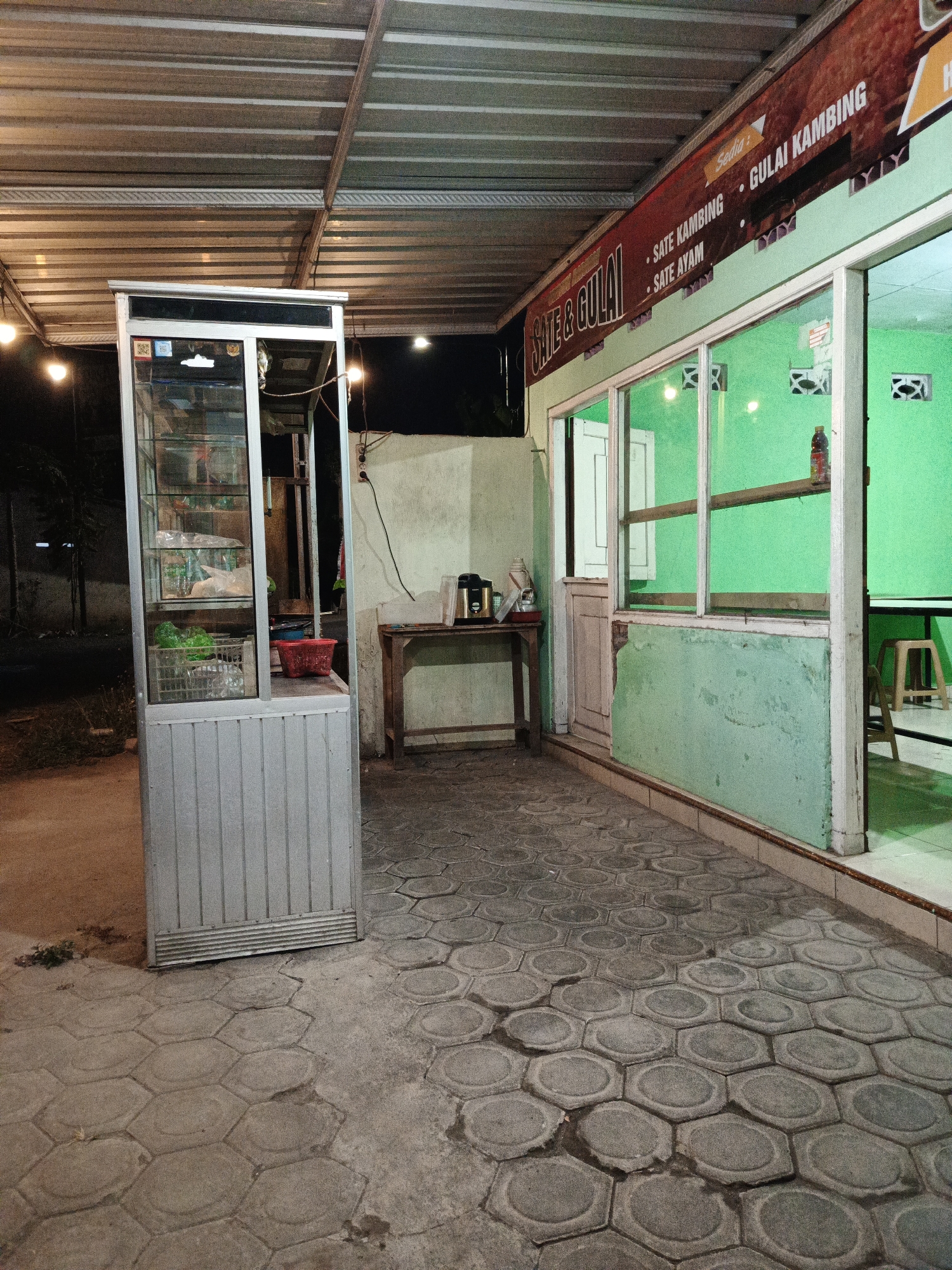 The image shows an outdoor food stall in Sumbersari, Indonesia, at night. The stall has a metal roof supported by metal beams. A glass display case, likely for food items, is positioned on the left side of the image. In front of the stall, a hexagonal patterned pavement is visible. To the right, a building with a light green exterior wall and large windows is present. A banner with text reading "Sate & Gulai" and "SATE KAMBING, GULAI KAMBING, SATE AYAM" is attached to the building's facade. Inside the building, green walls are visible, and a table with a stool can be seen through the windows. The overall scene suggests a simple, locally operated eatery.
