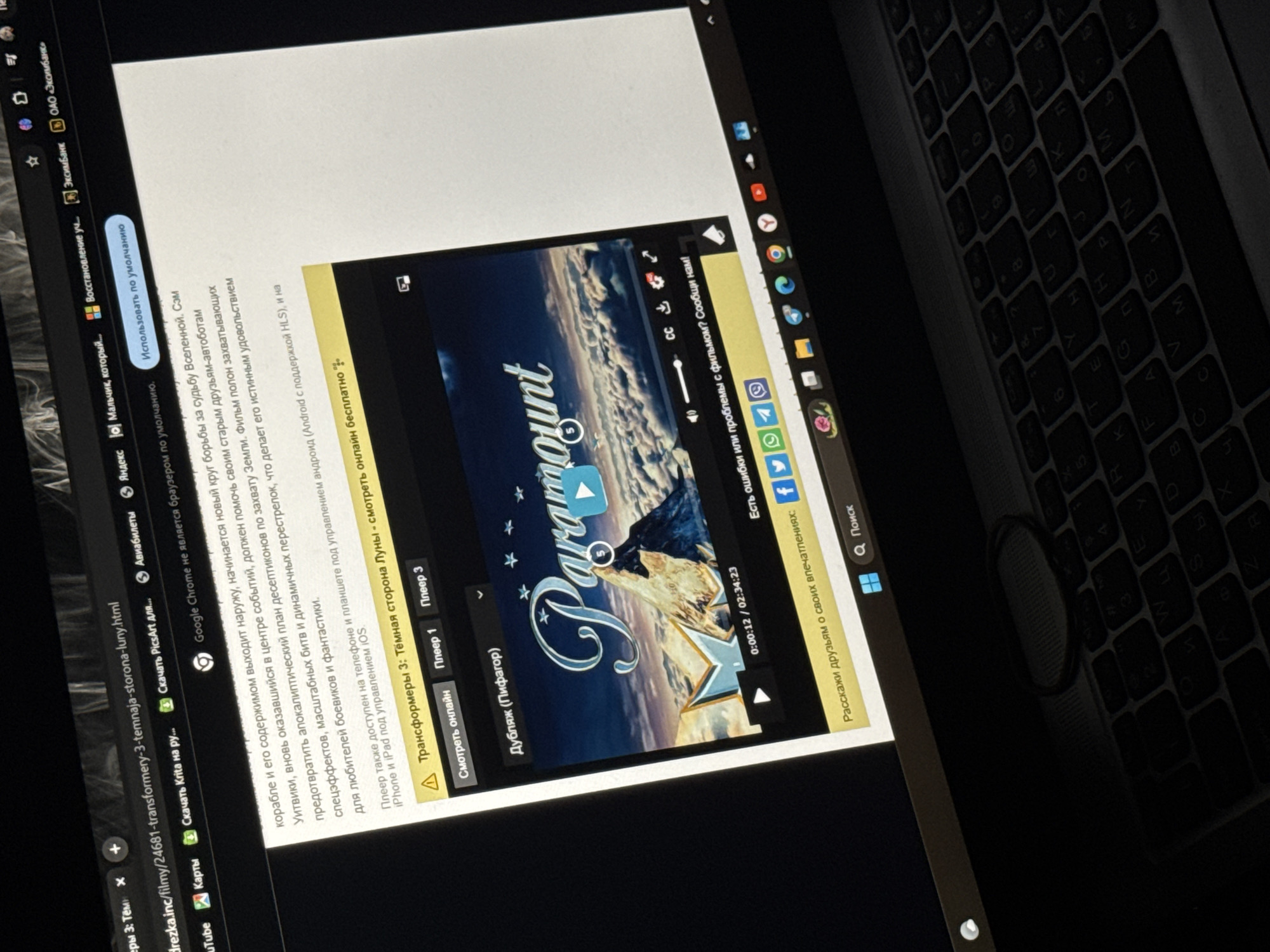 The image shows a close-up of a laptop screen displaying a webpage with a video player. The video is for "Transformers 3: Dark of the Moon" and appears to be playing. The browser tab indicates the URL is related to transformer-3-temnaja-storona-luny.html. The surrounding webpage content is in Russian and discusses the movie. The keyboard of the laptop is visible in the foreground, and a rubber band is resting on it. The scene is indoors, likely in a home or office environment, with no discernible external environmental features. There are no people visible in the image. The lighting appears to be from the laptop screen itself, with some ambient light contributing to the overall illumination. The image does not provide any information about the location in Tiraspol, Moldova.