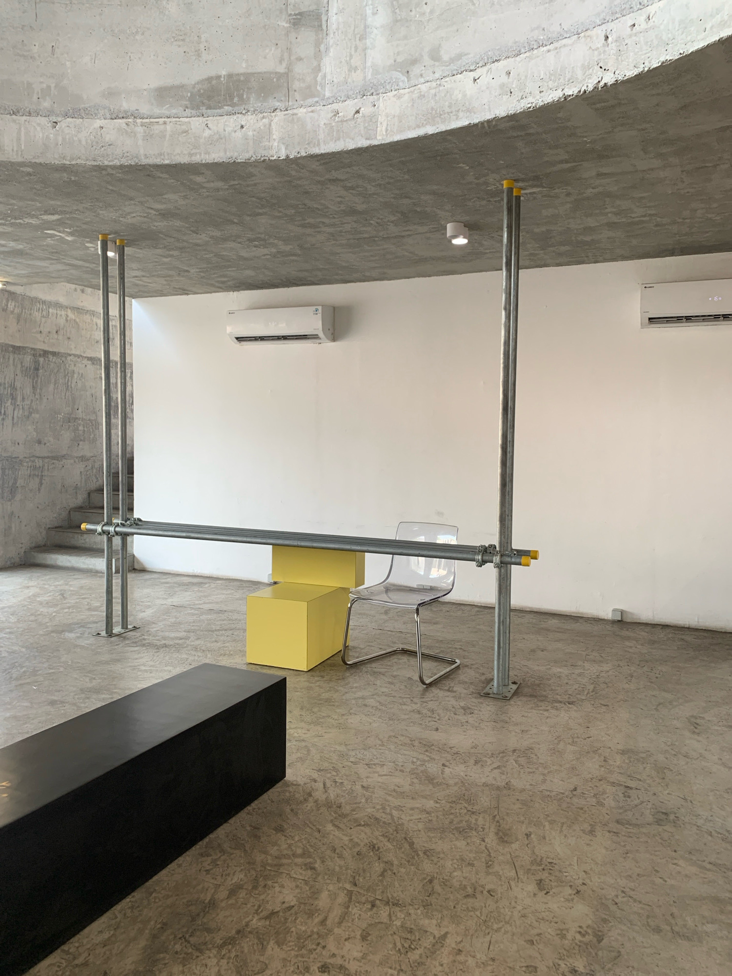 The image depicts an indoor space with a raw, industrial aesthetic. The walls are a mix of textured concrete and smooth white paint. The ceiling is a curved concrete structure. On the white wall, two modern air conditioning units are visible. In the center of the space, a horizontal metal pipe structure is supported by two vertical metal poles, resembling scaffolding. To the right of this structure, a clear acrylic chair with a metal frame is placed near a bright yellow cube-shaped block, which appears to serve as a low table or seating element. In the foreground, a long, black rectangular object, possibly a bench or a plinth, rests on the polished concrete floor. To the left, a concrete staircase with a rough texture leads upwards. The overall lighting is neutral, suggesting an interior space without direct sunlight. The location is identified as Meri, Indonesia.