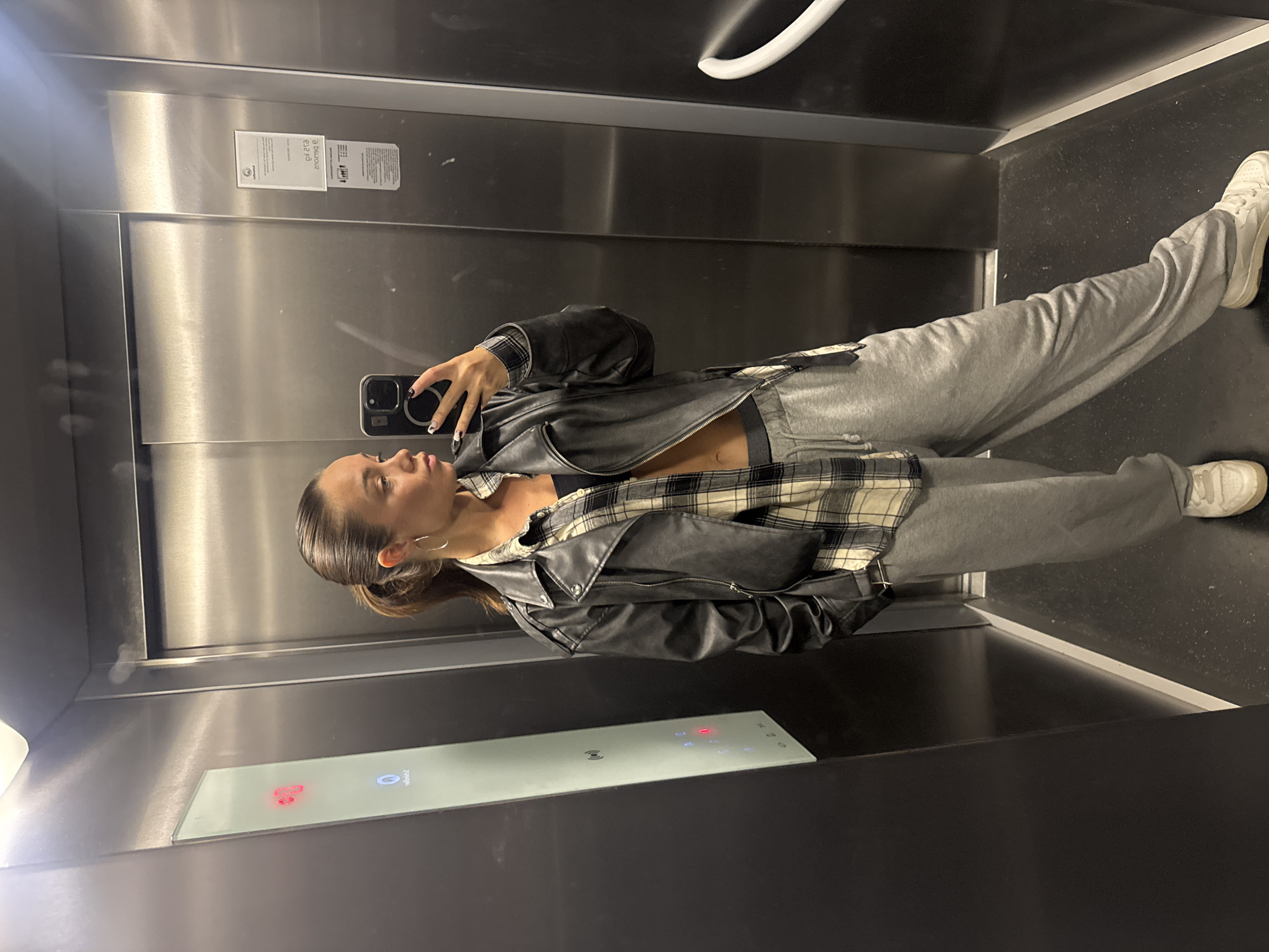 A young woman takes a selfie in an elevator with stainless steel walls. She is wearing gray sweatpants, white sneakers, a black sports bra, a plaid button-down shirt, and a black leather jacket. Her dark hair is pulled back, and she is wearing hoop earrings. The elevator control panel is visible in the lower center of the frame. Based on metadata, the location is identified as Budva, Montenegro.