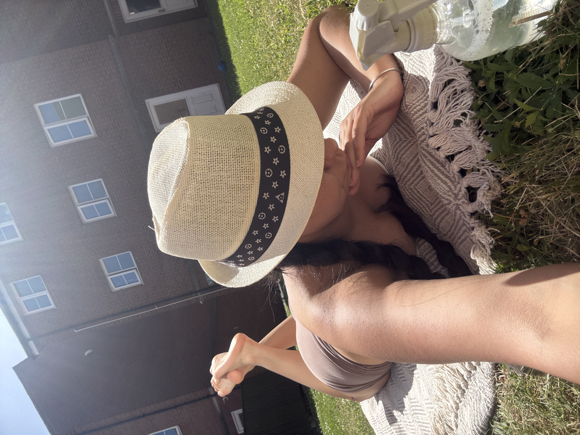 A woman is lying on a blanket in the grass, wearing a straw hat with a patterned band and a tan-colored bikini top and shorts. Her hair is braided and she is holding her hand to her face. A spray bottle sits next to her on the blanket, which is fringed. The location appears to be a yard with green grass, and a building with multiple windows is in the background. The image was taken in Great Yarmouth, United Kingdom.