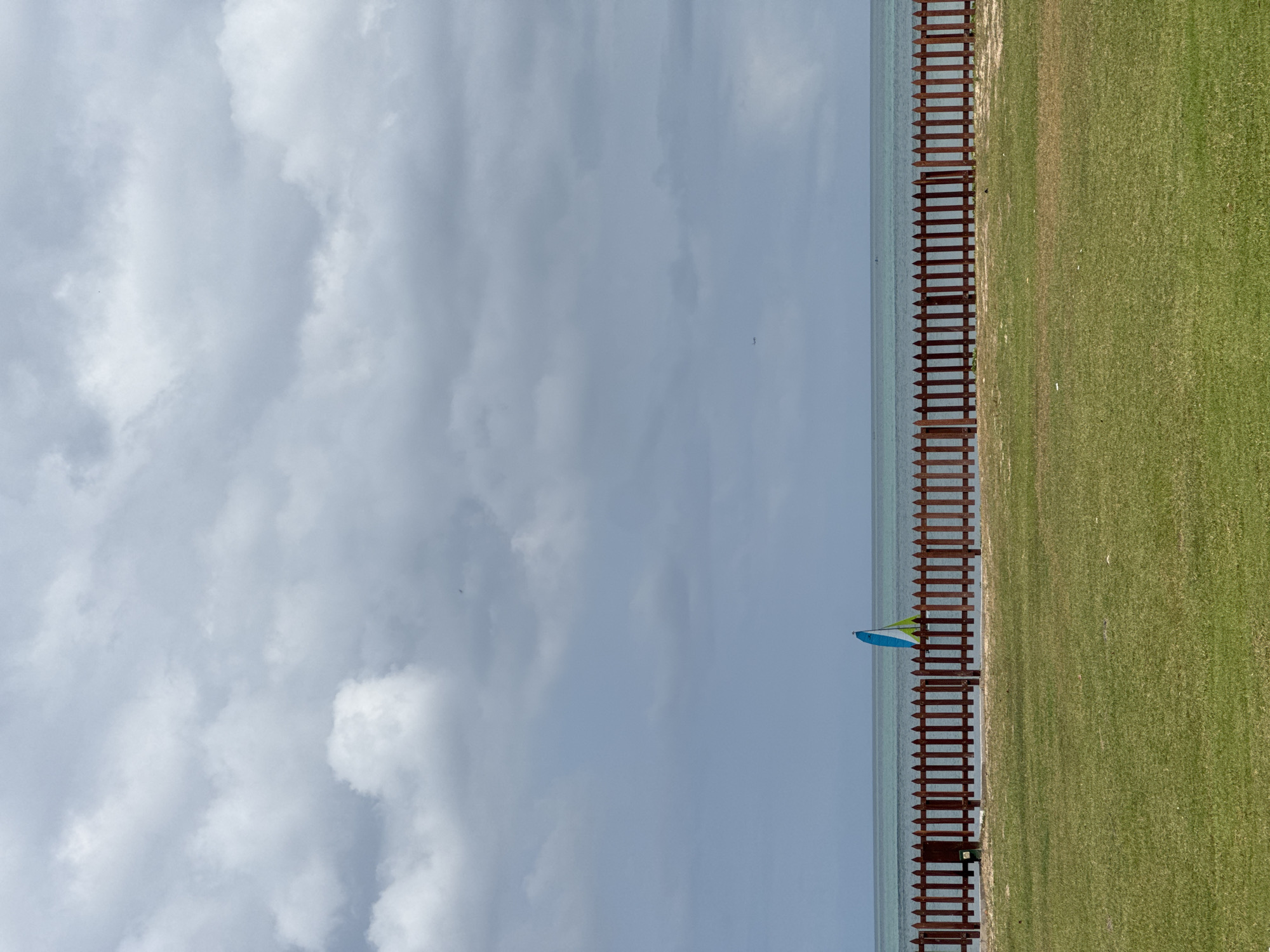 The image depicts a serene coastal scene under a cloudy sky. In the foreground, a meticulously maintained green lawn stretches towards a wooden picket fence. Beyond the fence, the tranquil turquoise ocean is visible, with a single sailboat featuring a green and blue sail gliding across the water. The horizon is clearly defined, separating the sea from the overcast sky. The overall impression is one of peacefulness and natural beauty. The scene does not provide any specific clues to pinpoint its location within Kuta Selatan, Indonesia.