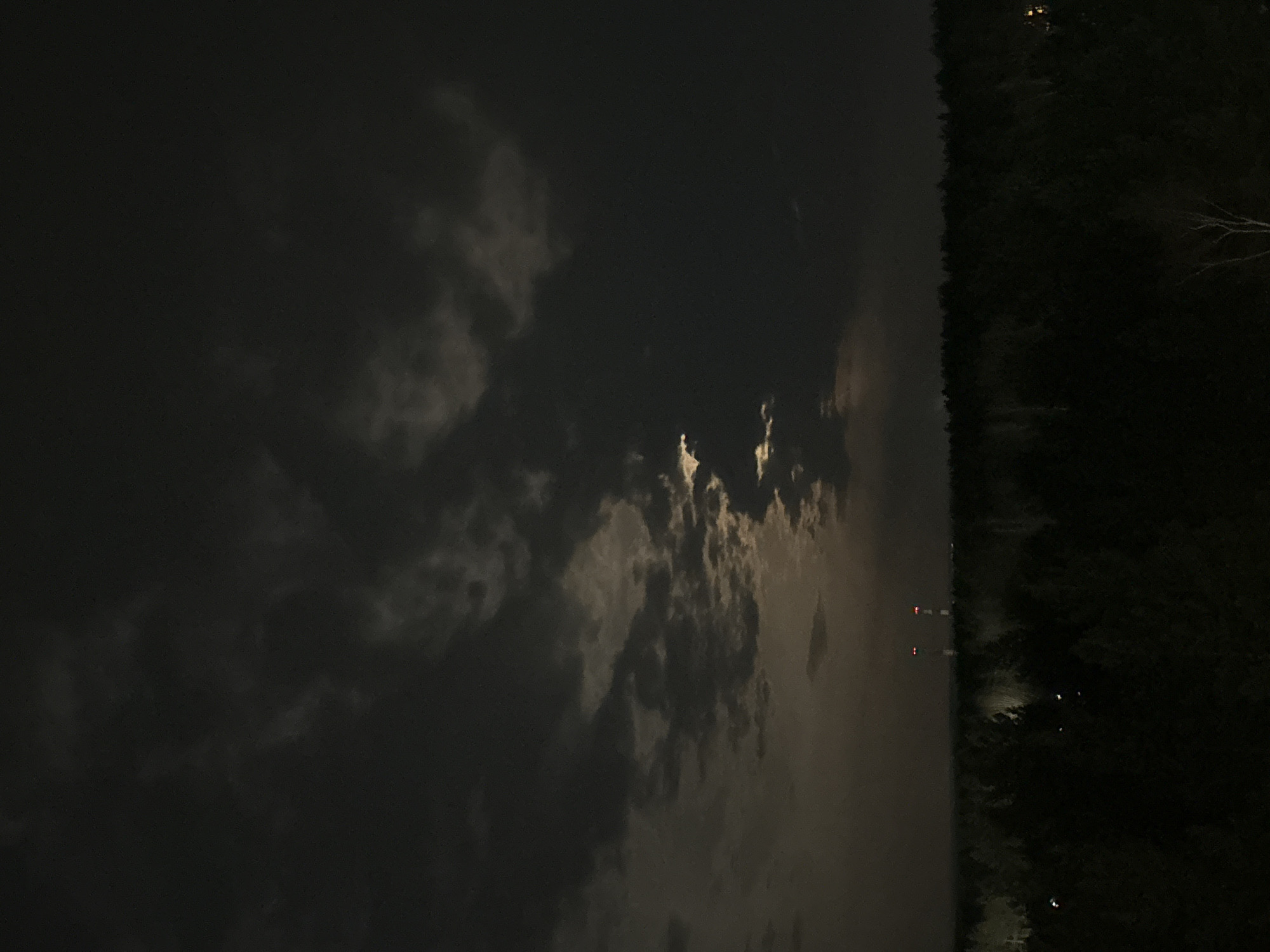 A nighttime photograph depicts a dark, cloudy sky over a forested area. The forest occupies the right side of the image, appearing dense and dark, with some illuminated spots that might be distant lights or residential buildings. Two tall structures with red lights are visible near the center of the horizon between the forest and the sky. The sky is filled with thick, dark clouds illuminated by the moon. The location is likely Chaikovsky, Russia, based on the presence of tall structures with red lights which may be associated with industrial areas or communication towers near forests.