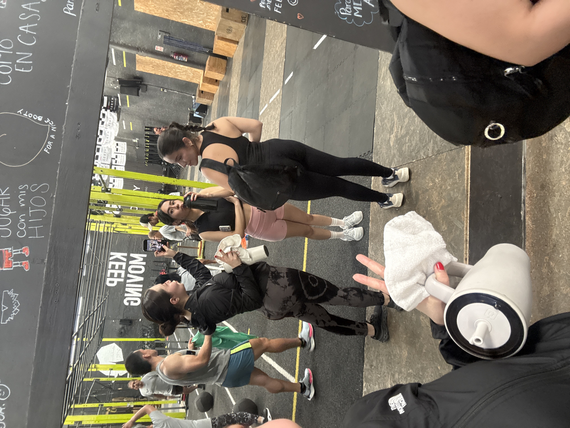 This image depicts an indoor group fitness class or training session in a gym or fitness studio, likely located in Mexico City as suggested by the visible Spanish text. The setting features a spacious area with black rubber flooring, concrete sections, and yellow demarcation lines, typical of a functional fitness or CrossFit-style facility. Overhead, a structure of metal racks and rigging is visible, along with wooden plyometric boxes in the background, one marked "30".
Multiple individuals are present, dressed in various athletic wear including leggings, shorts, sports bras, and sneakers. In the foreground, a person holds a white insulated water bottle and a small white towel, with red nail polish visible on their hand. Another woman in dark athletic wear, including a black top and leggings, carries a black backpack. Adjacent to her, another woman in a black top and pink shorts holds a dark water bottle. Further back, a person captures the scene with a smartphone. Men are also visible among the participants, including one in green shorts and a grey tank top.
The black walls are adorned with white handwritten text and drawings. Visible phrases include "JUGAR con mis HIJOS" (Play with my children), "COMO EN CASA" (Like at home), and a partially visible "FOR A NICE BOOTY" drawn around an outline. Mirrored text "KEEV ЗИVОМ" (likely "KEEP MOVING") is also present. White banners in the background display the text "XORYH". A person in the foreground wears a jacket featuring "THE NORTH FACE" logo. The overall atmosphere appears focused and active, characteristic of a workout environment.