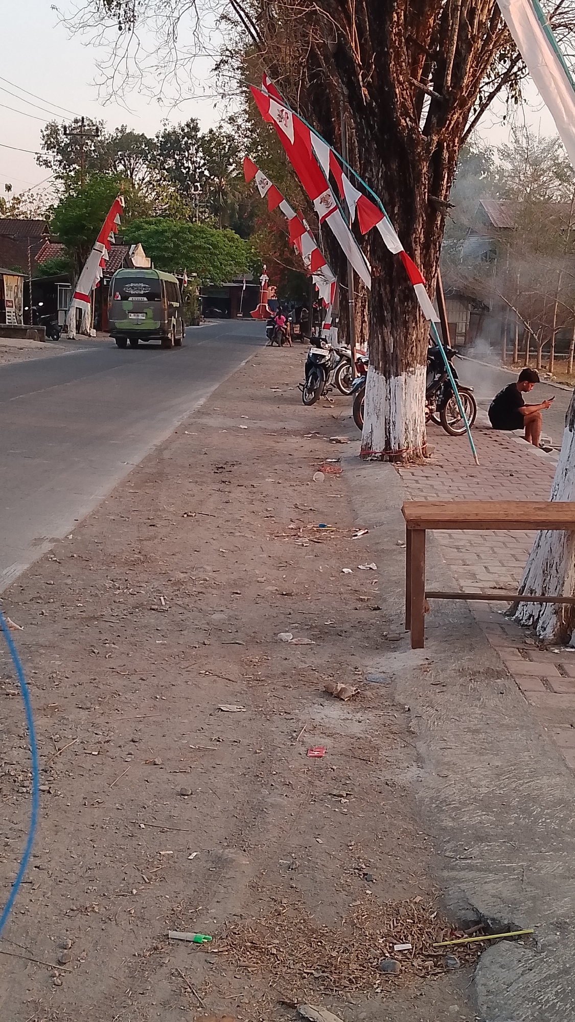 The image depicts a roadside scene in Karanglangu, Indonesia, characterized by a slightly dusty and littered ground. A paved road runs through the scene, with a dirt shoulder. Several Indonesian flags, red and white, are prominently displayed, strung along poles or trees, suggesting a festive or patriotic occasion.
In the background, a green minibus is visible on the road, moving away from the camera. Several motorcycles are parked along the side of the road. A man, dressed in a black t-shirt and shorts, is seated on a bench on the right side of the image, looking at his phone. The bench is made of wood and has a simple design. Trees line the road, with their trunks partially painted white at the base. The overall lighting suggests it is either early morning or late afternoon, with a warm, soft glow. The environment appears to be a mix of rural and semi-urban, with buildings visible in the distance.