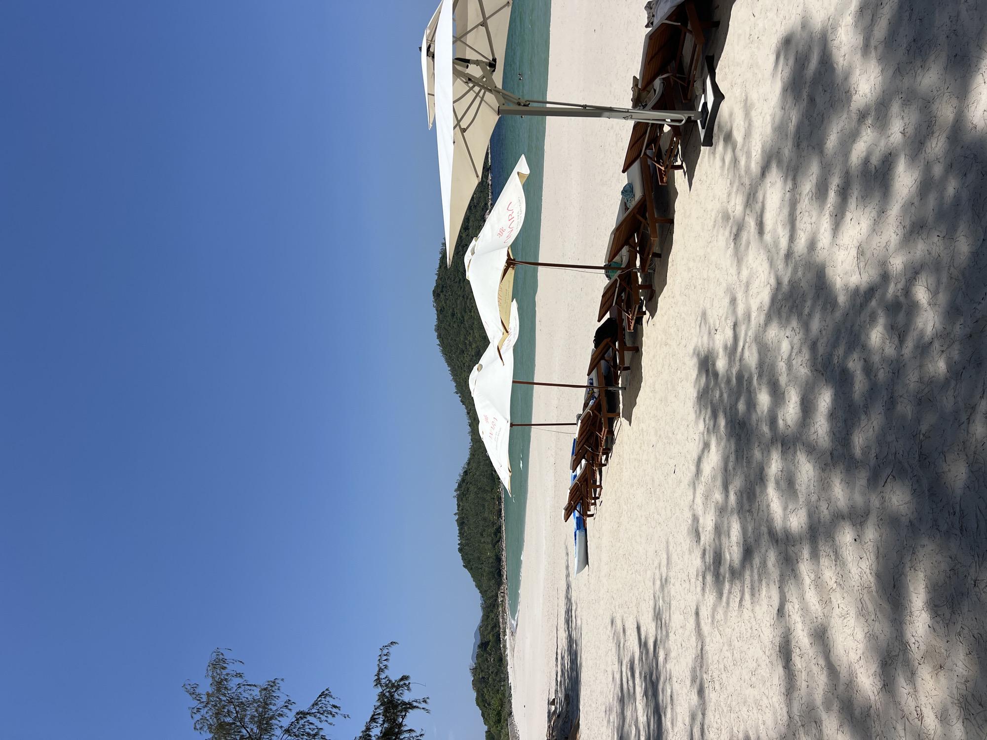 The scene captures an unoccupied white sand beach in Cong Hai, Vietnam, under a clear, cloudless blue sky. A series of wooden lounge chairs with light cushions and several white beach umbrellas are arranged linearly on the sand, with one umbrella displaying the text 'ANARA'. A white kayak or small boat is positioned on the sand adjacent to the chairs. Prominent shadows from overhead trees and umbrellas are cast across the foreground and mid-ground sand. The white sand beach extends into a calm bay featuring clear turquoise water, with verdant, tree-covered hills forming the distant backdrop. No individuals are visibly present.