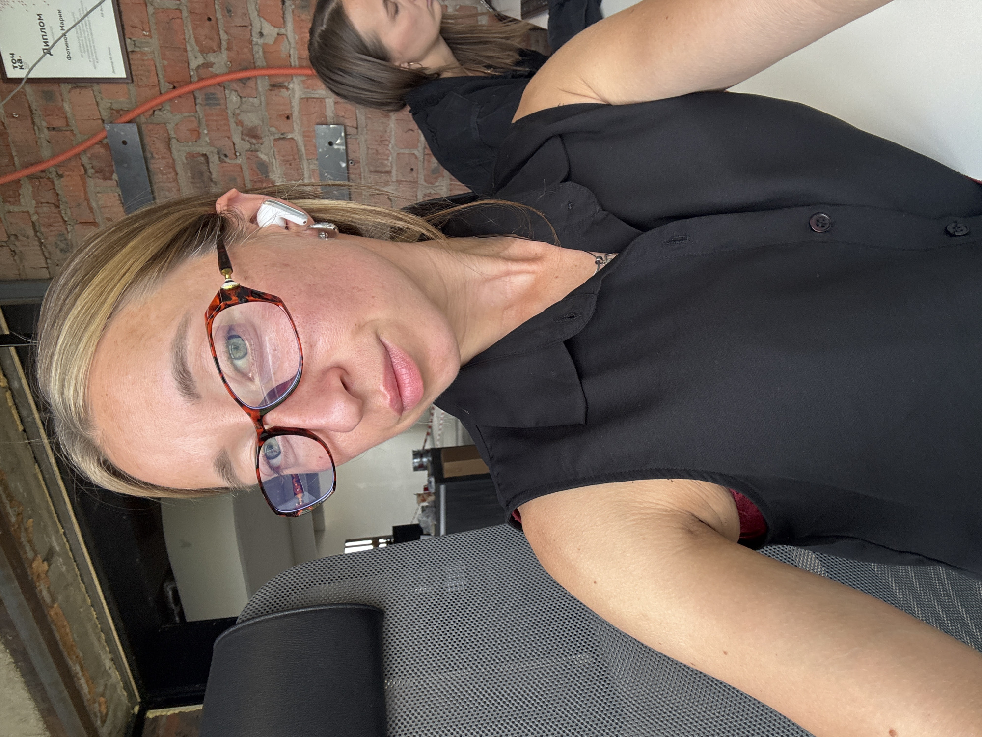 This is a vertical, close-up photograph of two women. The woman in the foreground is looking towards the camera and smiling. She has blonde hair pulled back, is wearing a black shirt, glasses with tortoise-shell frames, and wireless earbuds. The second woman is in the background, with her head resting on her arm, appearing to be asleep or resting. She has long brown hair and is also wearing a black shirt. The setting appears to be indoors, with a brick wall and a framed certificate visible in the background. The overall lighting suggests it is daytime. There is some text visible on the certificate in the background, appearing to be in Russian.