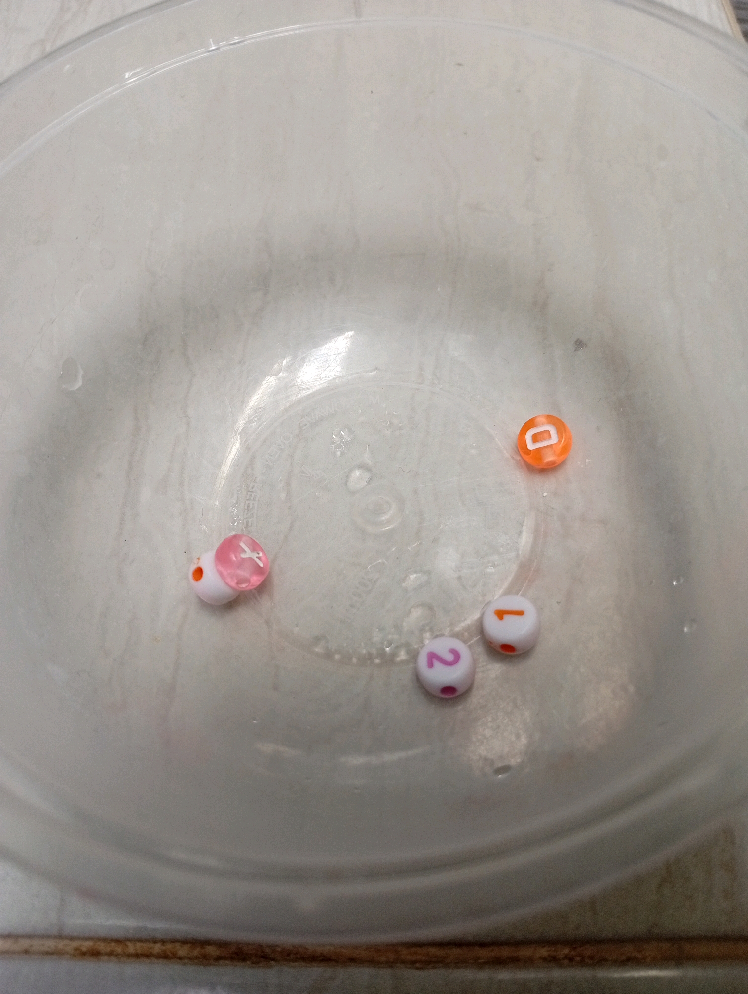 A clear plastic bowl rests on a light-colored, textured surface. Inside the bowl are five small beads. One bead is orange and has the letter "D" on it. Three beads are white with pink dots around the edge; one has the number "1" on it, another has the number "2" on it, and the third is blank. The fifth bead is pink and clear with a white "X" on it, and it is paired with a small white bead. The scene is well-lit, and the focus is on the beads within the bowl. There is no indication of the city of Kebondalem, Indonesia, or any people present in the image.