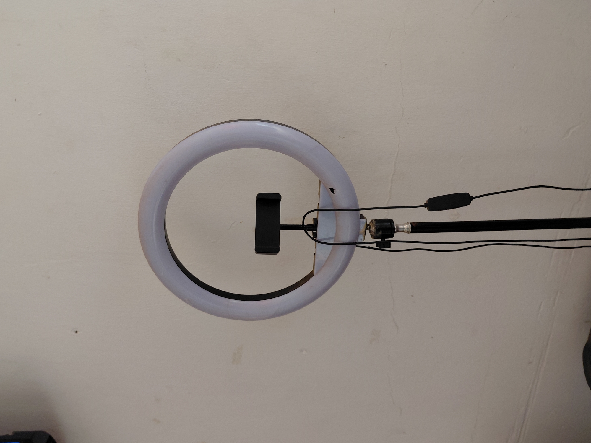 The image displays a white ring light, oriented horizontally, against a light-colored, textured wall. A black phone holder is mounted centrally within the ring light's circumference. From the right side of the ring light, multiple black cables extend, one of which incorporates an oblong black inline switch or controller. These cables are positioned alongside a black pole or stand, which supports the ring light assembly and extends further to the right.
The background consists of a plain, off-white or light beige wall that shows signs of wear, including subtle surface textures, fine cracks, and minor smudges or discolorations. No people are present in the scene, and no specific activity is taking place. There is no discernible text visible anywhere in the image. The setting appears to be an indoor space.