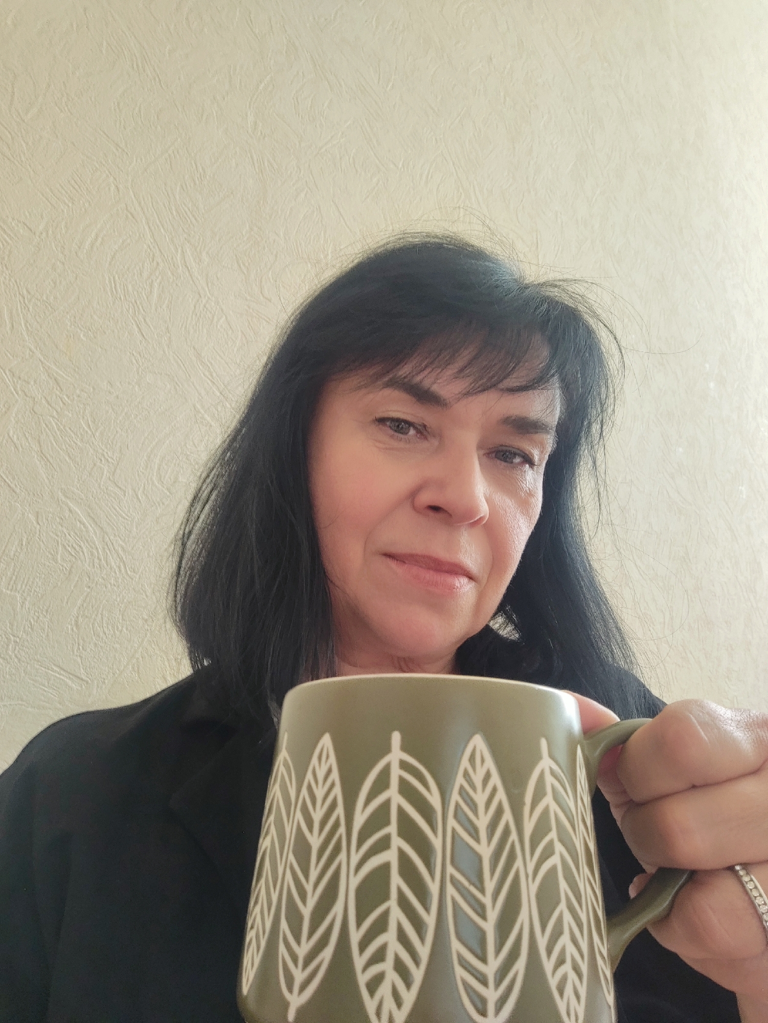 A medium shot captures a woman holding a green mug with a leaf pattern. She has dark, shoulder-length hair with bangs and appears to be in her 50s. The woman is looking towards the camera with a neutral expression. She is wearing a black blazer. The background is a textured, light-colored wall, suggesting an indoor setting. The lighting is soft and diffused, indicating it could be daytime, but the exact time of day and weather conditions are not discernible. There is no visible text in the image.