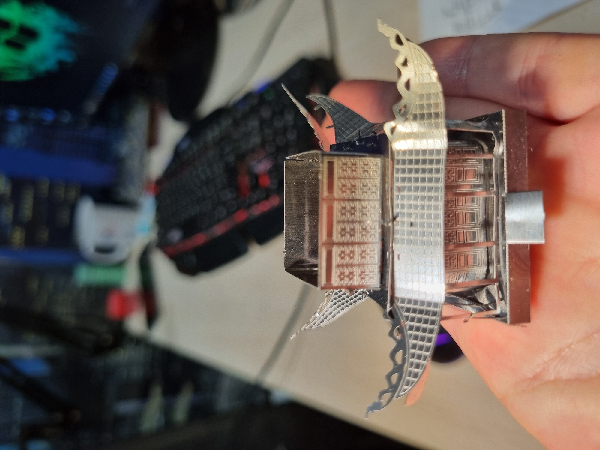 A close-up shot shows a hand holding a partially assembled, intricate metal model resembling a pagoda or temple. The model features detailed facades with repetitive geometric patterns, decorative archways, and possibly a spire or finial. The model is made of thin, polished metal. In the background, slightly out of focus, there is a keyboard, part of a computer, and a desk. The location is not specified but there is no information provided in the image that identifies its location as Chaikovsky, Russia.