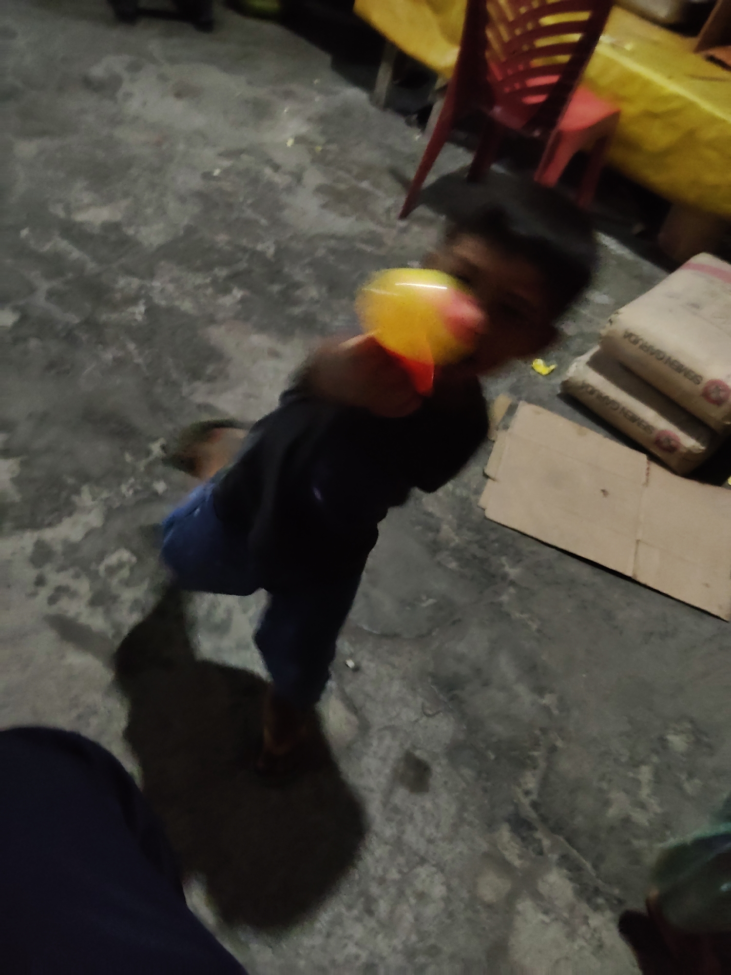 A child is playing with a toy in a cluttered outdoor area, possibly a yard or an alley, in Mekarmukti, Indonesia. The child, wearing a dark long-sleeved shirt and blue pants, is holding a colorful toy, possibly a rattle or ball, in their right hand. The toy is blurred due to movement, as is the child's face, suggesting a candid moment. The ground is a rough, gray concrete surface with some scattered debris and a dark shadow cast by the child. In the background, there are stacks of cement bags, a red plastic chair, and yellow sheeting, indicating a construction or storage area. The overall lighting suggests it is either evening or a dimly lit area.
