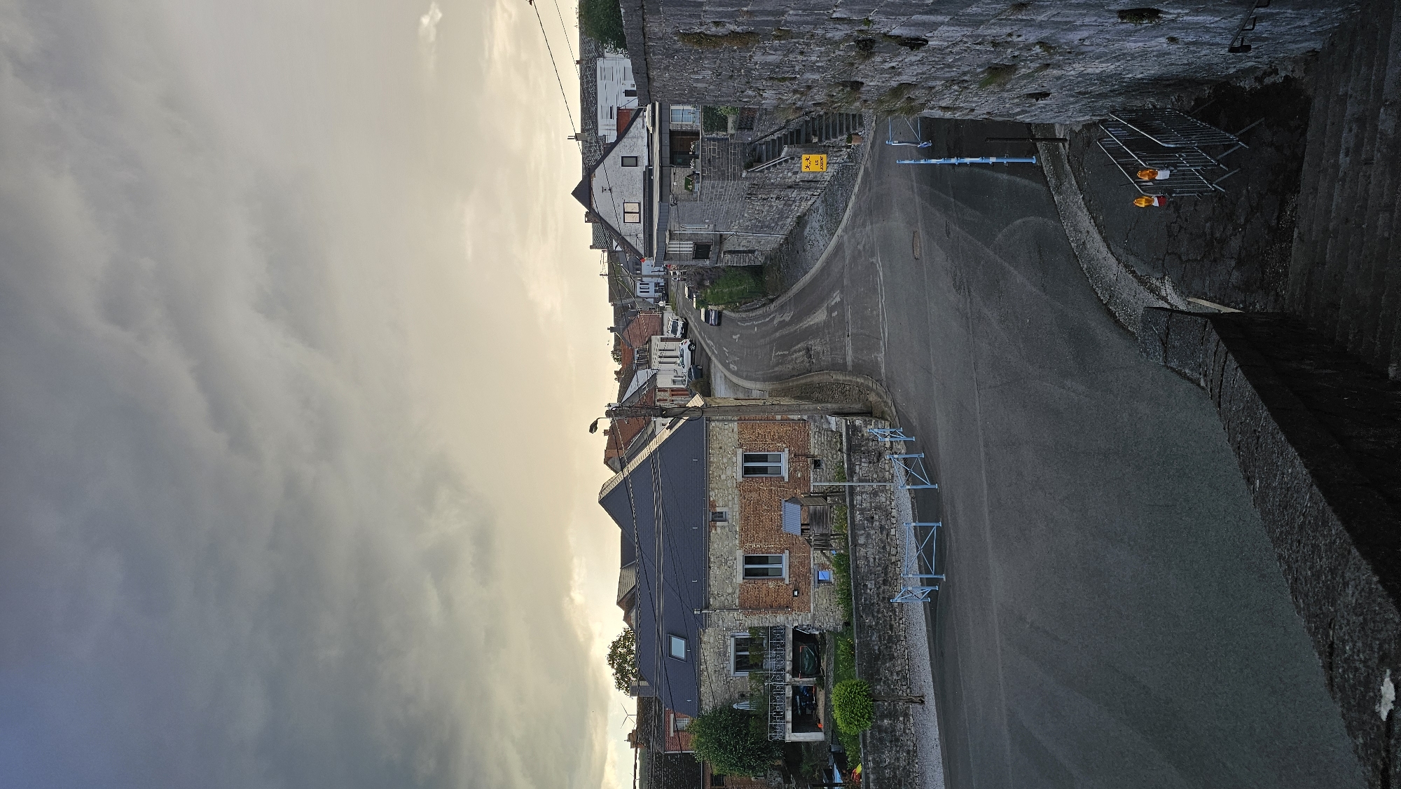 The image displays a quiet, winding street in the Belgian village of Boussu-lez-Walcourt under an overcast sky, suggesting either early morning or late afternoon. The sky is largely covered with thick, grey clouds, though a subtle warm, golden light appears on the right side, possibly from the sun breaking through or setting.
The asphalt road curves gently, flanked by traditional European village architecture. Buildings are constructed primarily from stone and brick, featuring varying roof types, including dark grey and red tiles. Stone retaining walls line both sides of the street, with a particularly prominent one on the right, which also incorporates a set of stone steps leading upward.
A few parked vehicles are visible further along the street. Streetlights and overhead utility lines are present. In the foreground on the left, a notable building with a dark grey roof and a facade of exposed brick and stone features windows with blue frames. On the right, metallic construction barriers with orange warning lights are visible next to the road, along with some lighter blue metal barriers. A yellow, diamond-shaped road sign on the right bank contains partially legible text, appearing to say "ATTENTION RALENTIR," indicating a warning to slow down.
The scene is devoid of people or direct activity, lending it a tranquil and possibly melancholic atmosphere.