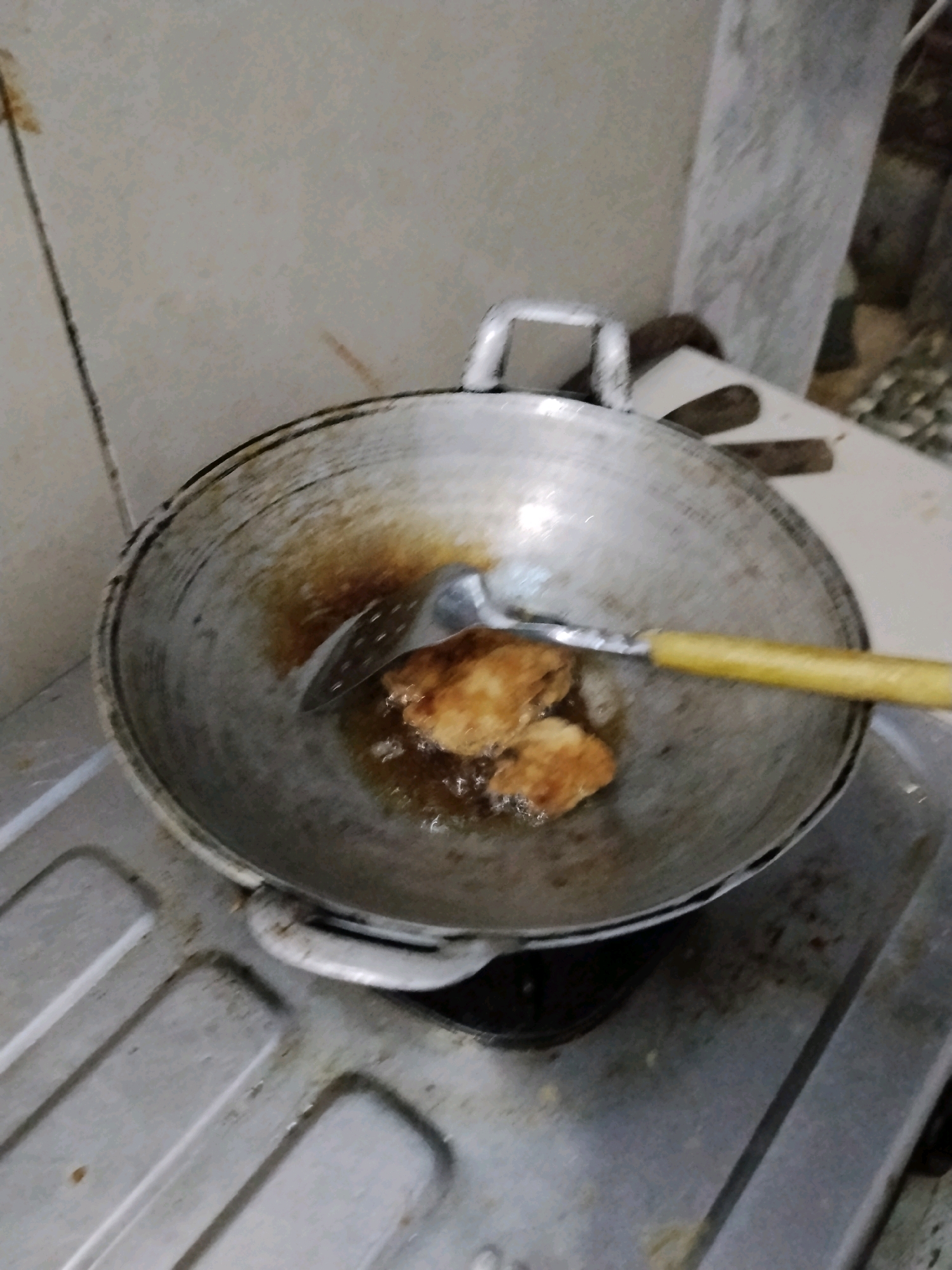 In a domestic kitchen setting, possibly in Kejawan, Indonesia, a close-up, slightly angled shot captures pieces of food being fried in a well-worn metal wok. The wok, showing signs of use with dark residue, rests on a silver gas stove. Two golden-brown food items, appearing to be pieces of fried chicken or fish, are submerged in sizzling oil. A metal slotted spatula with a yellow handle is positioned to lift or turn the food. The background consists of a light-colored tiled wall, showing some staining, and a blurred view of what appears to be a textured column or pillar. The lighting is dim, contributing to the intimate, everyday scene of cooking.