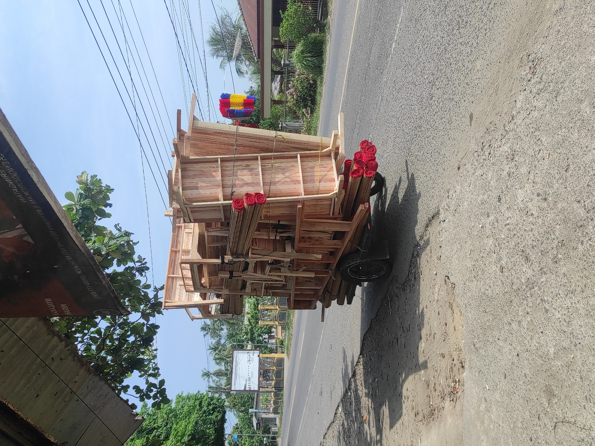 The image shows an overloaded two-wheeled cart on a road in Keudeteupinpunti, Indonesia. The cart is loaded with light-colored wood frames and wrapped red objects, possibly rugs or mats. A person is implied to be maneuvering the cart. The road runs alongside buildings and foliage. Overhead power lines are visible. A colorful, stacked, rectangular object is seen in the background.