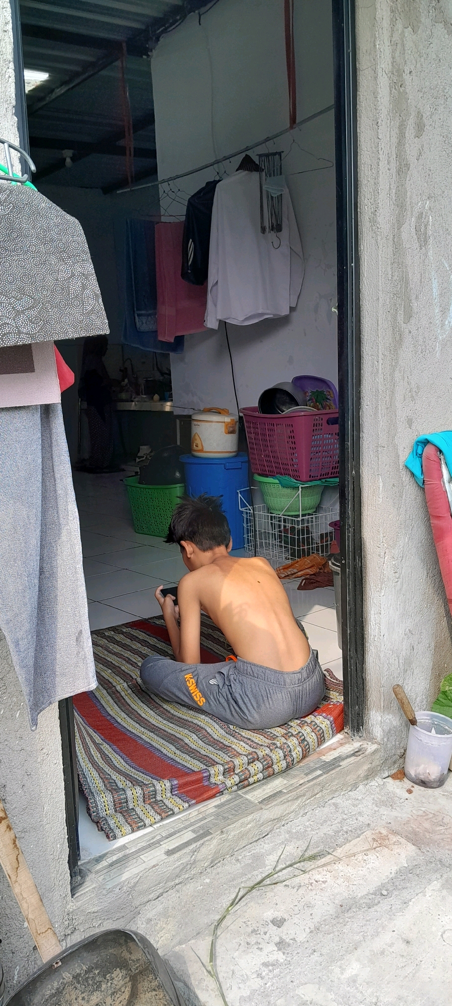 The image shows a young male, shirtless and wearing grey sweatpants, sitting on a colorful striped mat outside a doorway. He is looking down at a mobile device he is holding. Behind him, through the open doorway, a woman in dark clothing is visible near what appears to be a kitchen sink area. The environment suggests a modest living space, possibly in Rejosari, Indonesia, with laundry hanging on a line inside, baskets of items, and household appliances. The exterior wall is textured and weathered.