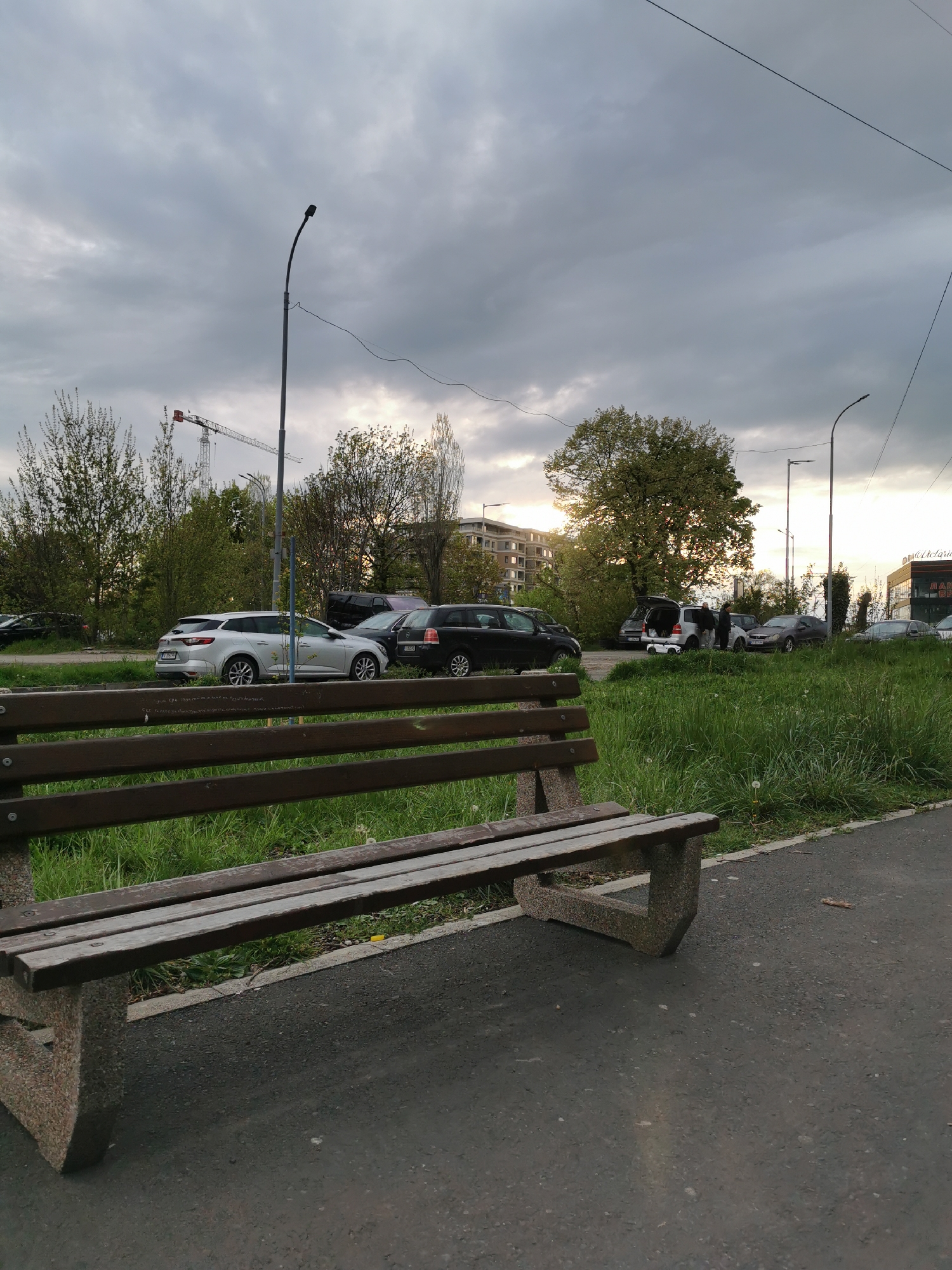 This media file depicts an overcast urban or suburban scene, likely in Burgas, Bulgaria, during late afternoon or early evening. In the foreground, a weathered public bench with dark wooden slats and rough concrete supports occupies the lower left corner, beside an asphalt path.
Beyond the path, a strip of unkempt green grass separates the foreground from a parking area. Several cars are parked on a dirt lot, including a silver station wagon (with a license plate partially visible as "B 188..." or "B 155..."), a black sedan, and a dark SUV/minivan. The trunks of some vehicles are open, suggesting activity such as loading or unloading. A few people are gathered around the parked cars, conversing or attending to items in the trunks; a small white toy car is visible on the ground near one open trunk.
The mid-ground features several trees with fresh spring foliage, some appearing denser than others. Tall, dark streetlights with overhead power lines are scattered throughout the scene. In the background on the left, a prominent red and white construction crane rises above the trees, indicating ongoing development. To the right, parts of multi-story buildings and a commercial establishment are visible. A sign on the building on the far right clearly shows the word "МАРКЕТ" (Market), with other letters possibly spelling "АПТЕКАРИ" (related to pharmacy). The sky is heavily clouded, creating a soft, diffused light across the landscape. The overall impression is a casual, everyday outdoor setting.