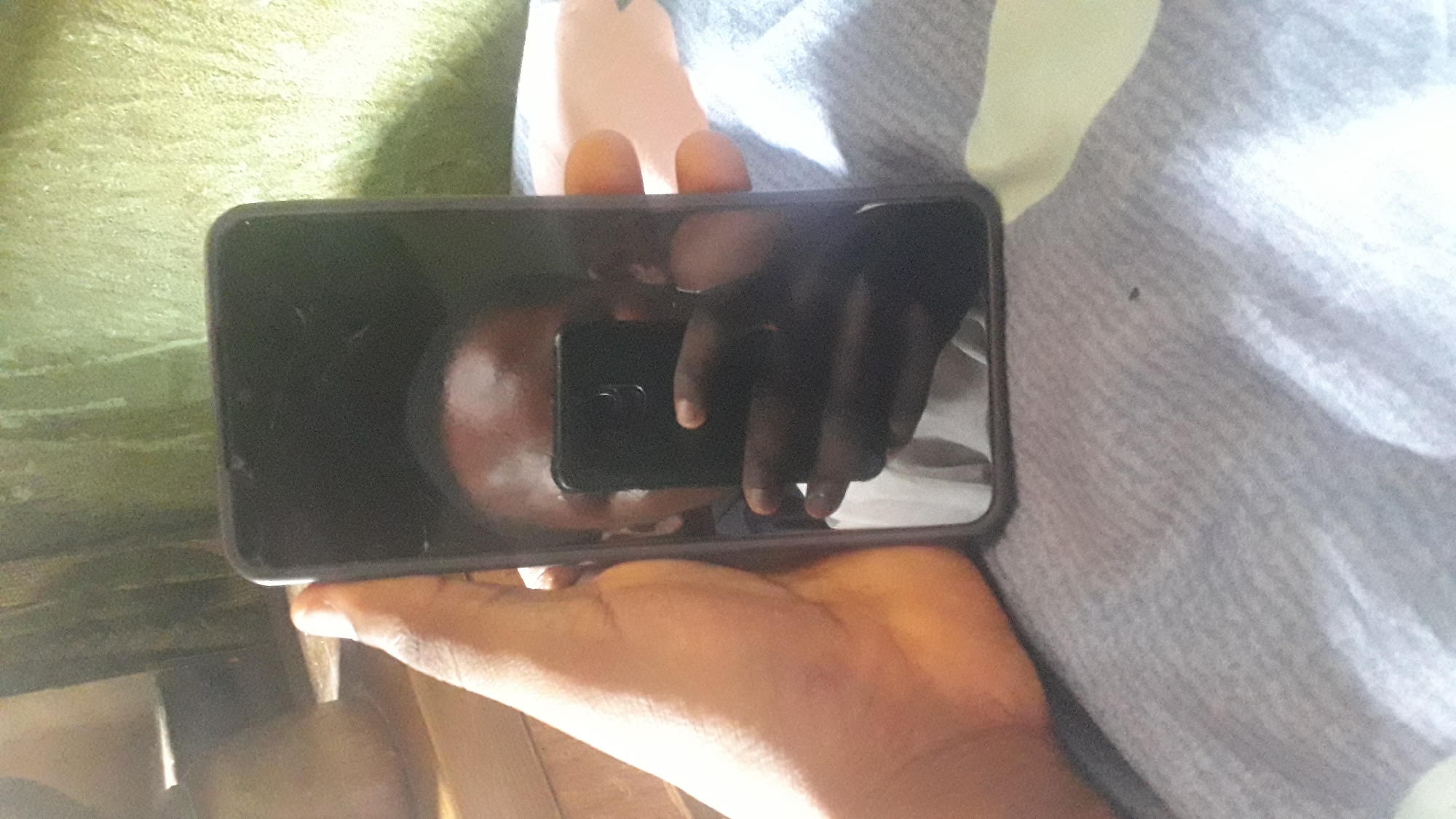 A medium shot shows a person holding a smartphone, which is capturing their reflection. The smartphone is black and held vertically by the right hand. The reflection in the screen shows the person's face, wearing a white shirt, and their hand holding the phone. The background is indistinct, but appears to be a wall covered with a green, textured material and what looks like a wooden surface on the left. The person is likely in Nkpor, Nigeria, based on context provided. The overall scene is dimly lit, with the focus on the smartphone and its reflection.