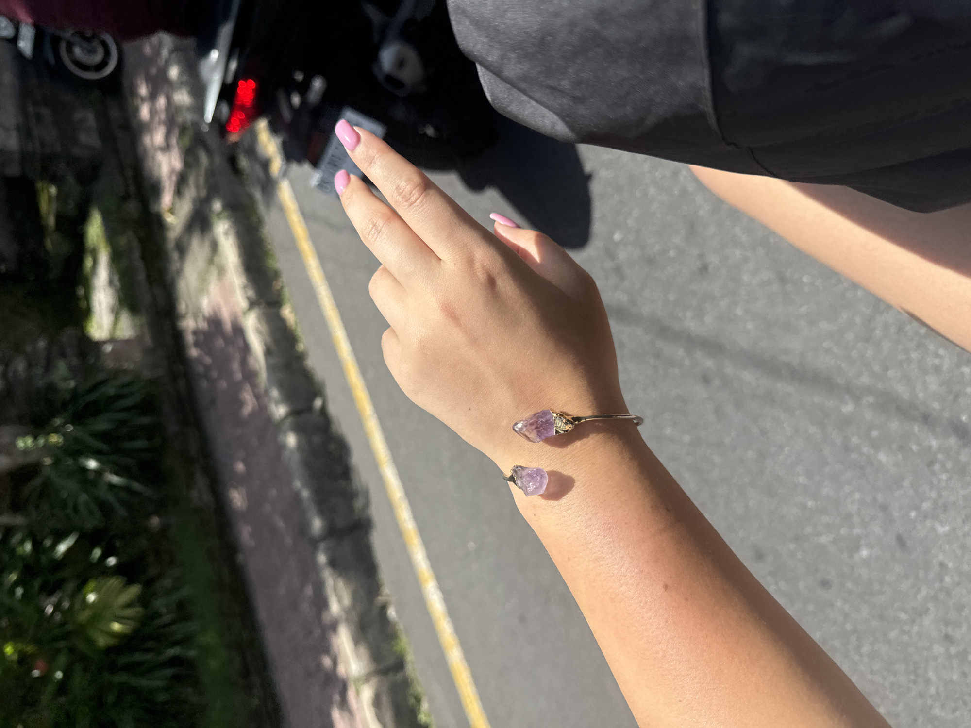 A woman’s arm and hand are shown wearing a cuff bracelet with purple gemstones. The nails are painted light pink. The woman is standing outside on a paved area. Beside her, there are plants and a vehicle. The location is Ubud District, Indonesia.