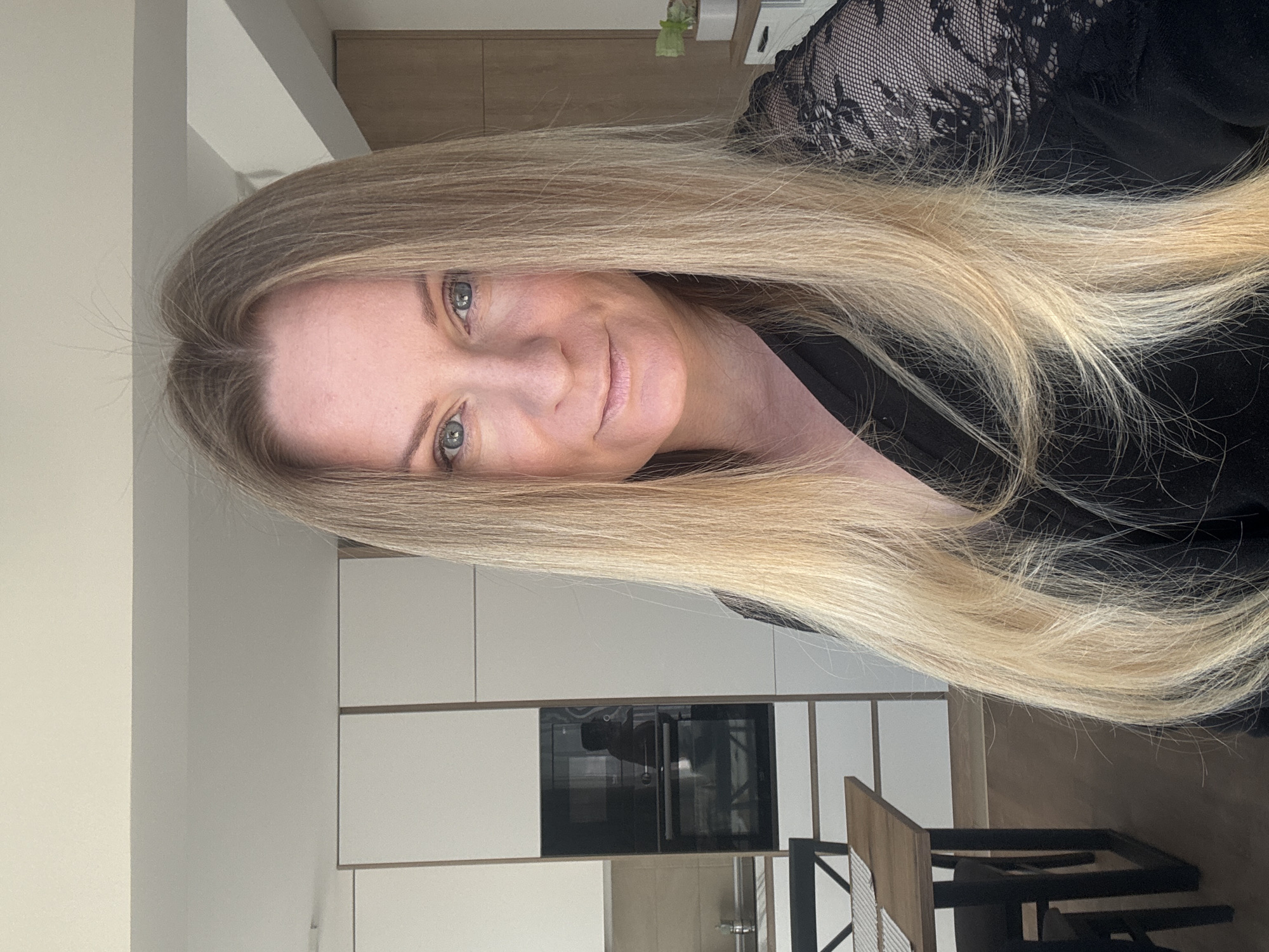 A blonde woman with light-colored eyes and a fair complexion is depicted in a medium close-up, posing for a photo. Her head is tilted slightly, and she has a soft, pleasant smile on her lips. Her long, straight blonde hair flows over her shoulders and chest. She is wearing a dark top, possibly black, with a visible lace pattern on her right shoulder/sleeve, which appears to be part of the garment.
The setting is indoors, appearing to be a modern kitchen or living area. The background features light-colored walls and contemporary white kitchen cabinets, some with a wooden accent on top. A built-in appliance, likely an oven or microwave, with a dark, reflective glass door, is visible in the cabinetry behind her. Below the cabinets, there's a dark floor, and to the right, a section of a wooden and metal table or bar counter with chairs can be seen. A small plant in a white pot is faintly visible on a shelf or surface in the upper right background. The lighting is soft and even, suggesting it is daytime indoors, though no direct windows or sunlight are apparent. There is no visible text in the image.
