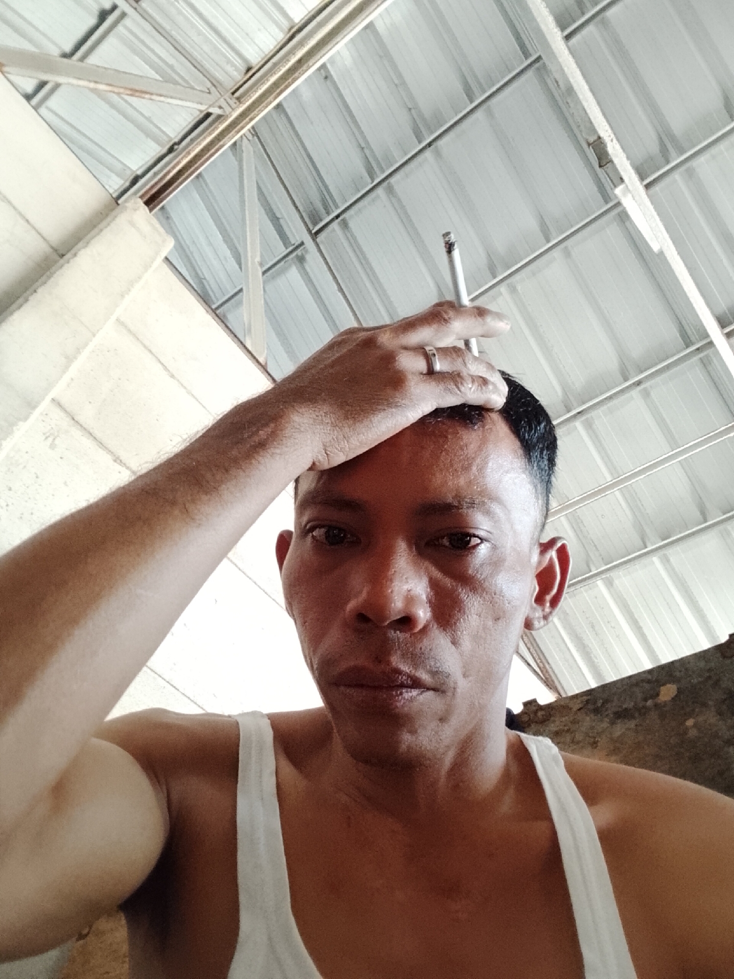 A low-angle, close-up, selfie-style shot captures a man in a white tank top as he places his right hand on his forehead. He is holding a cigarette between his right index and middle fingers, which is lit. The man has short, dark hair, and his face is turned towards the camera. A silver ring is visible on his ring finger. The background reveals a corrugated metal ceiling with exposed beams and a rough, concrete wall. The setting appears to be an indoor, possibly unfinished, structure.