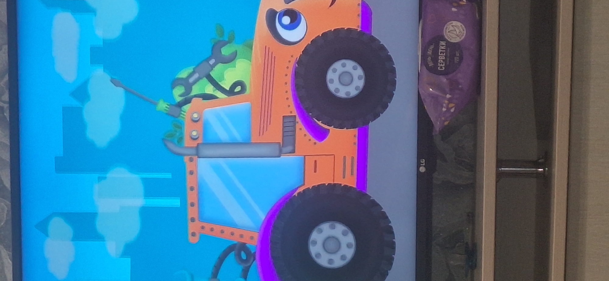 The image is a photograph of a television screen displaying a cartoon. The cartoon depicts an anthropomorphic orange tractor with large black wheels and purple fenders. The tractor has a smiling face with a large blue eye. It is set against a blue sky with cartoonish clouds and city building silhouettes. In the foreground, below the television screen, is a shelf with a package of wet wipes. The television itself has an LG logo on its lower bezel. The provided crops show close-ups of various elements of the cartoon and the lower portion of the television and shelf. There is no information within the image or its crops that indicates a specific location in Bila Tserkva, Ukraine.