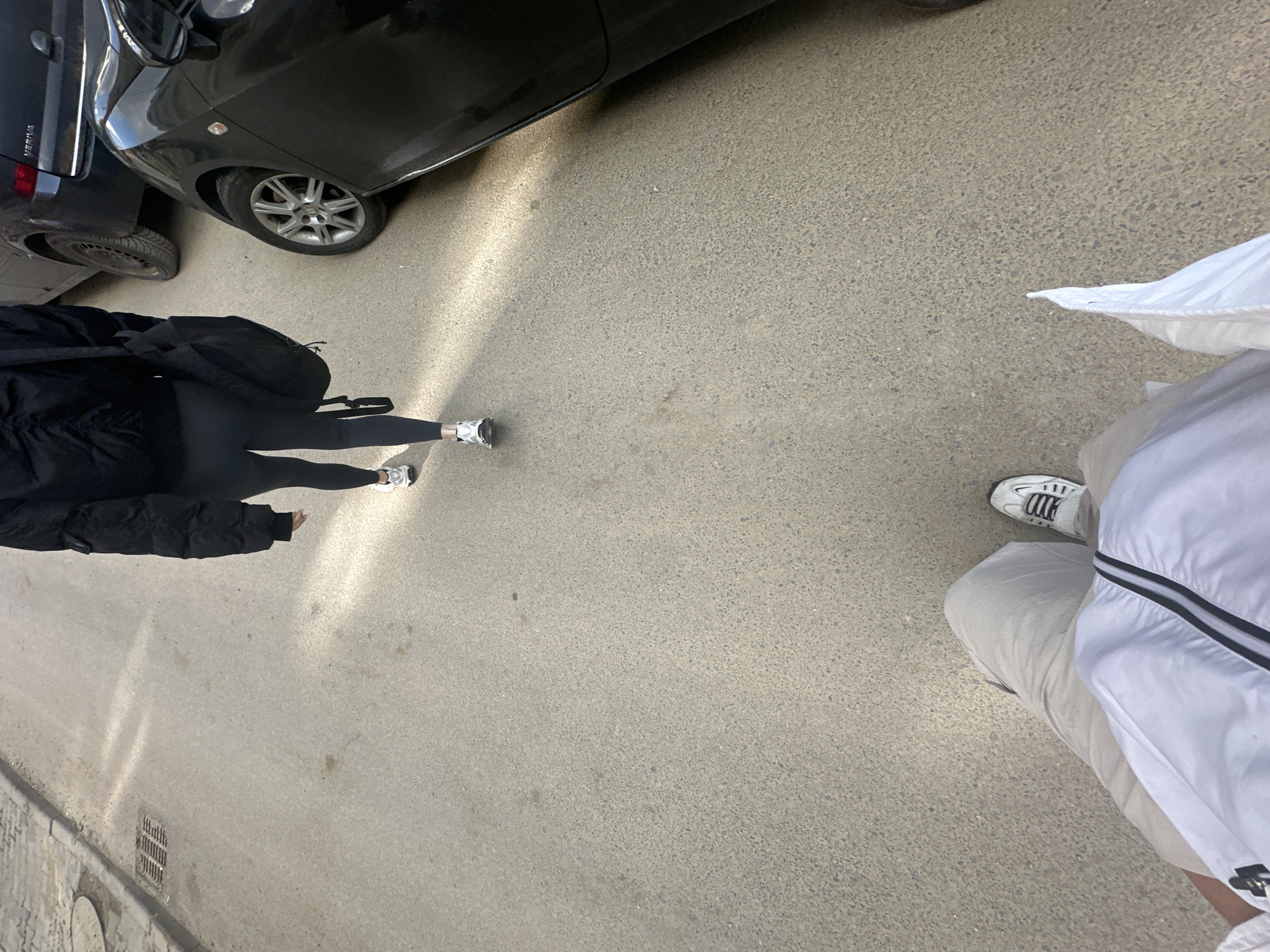 The image shows a street scene, possibly in Sofia, Bulgaria, though no specific landmarks are visible. Two individuals are partially in frame. One person is wearing a black jacket, black leggings, and white sneakers. A black bag is slung over their shoulder. Another person is wearing beige pants, white sneakers, and a white jacket with black stripes. Two cars are partially visible; a dark-colored car in the upper-left corner, and another darker colored car farther to the left. The ground is a light grey pavement.