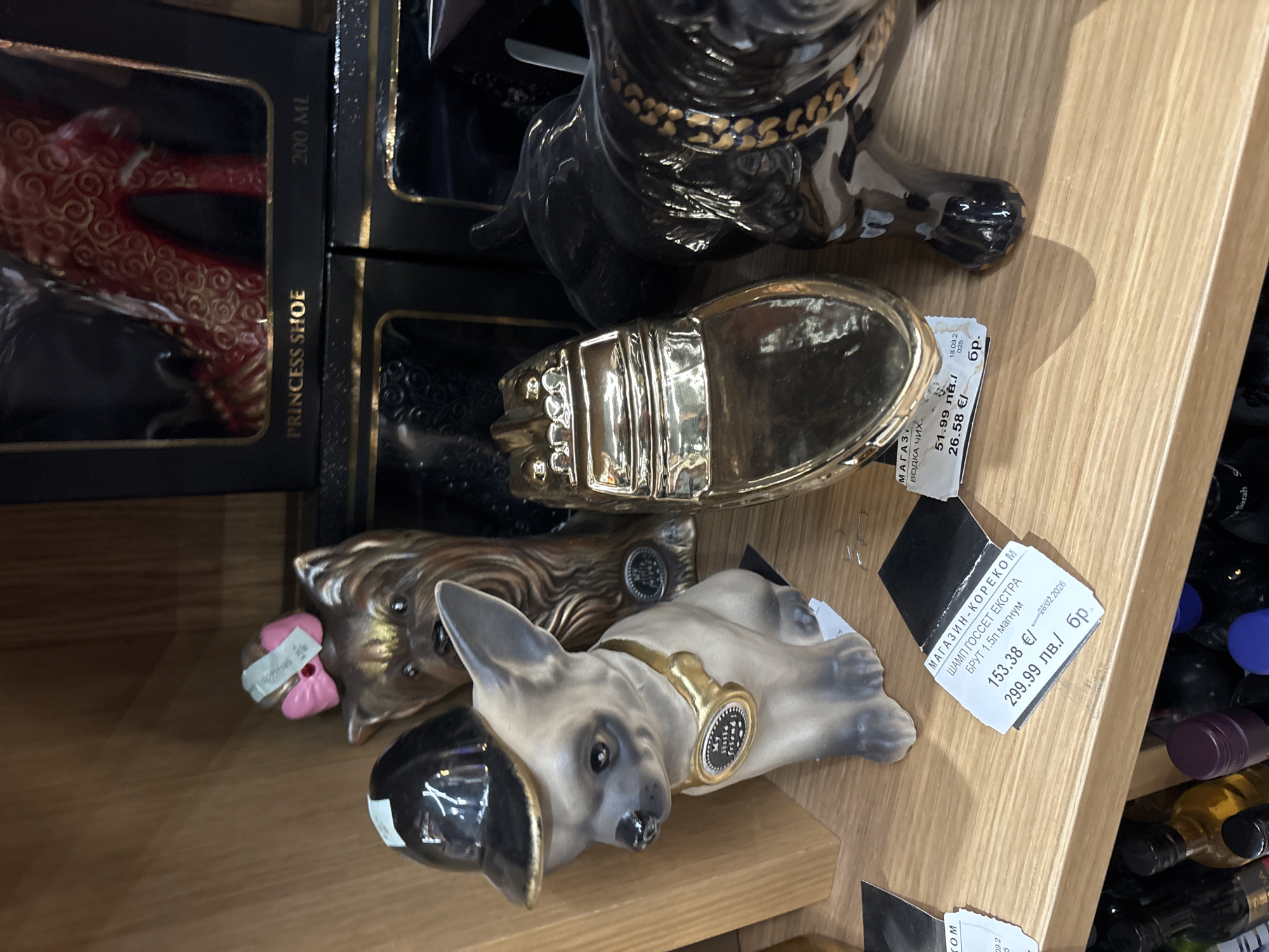 The image shows a shelf in a store, possibly in Plovdiv, Bulgaria. The shelf contains several decorative items: ceramic dog figurines, and "PRINCESS SHOE" packaging. Price tags are visible with prices in both Euro and Bulgarian Lev. Bottles of wine are visible in the background on the right.