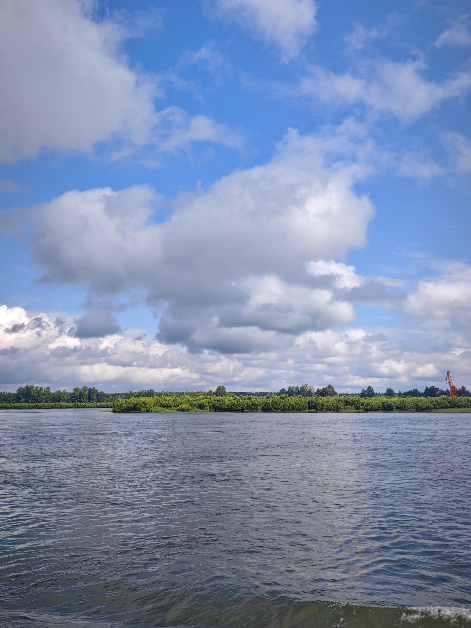 A wide, calm river dominates the foreground of this image. The water is a dark blue-gray and shows gentle ripples. In the distance, a lush, green treeline stretches across the horizon, suggesting a dense forest or vegetation. Above the treeline, a bright blue sky is filled with large, fluffy white clouds. On the far right side of the treeline, a tall, red crane is visible, indicating some form of industrial activity or construction. The scene appears to be a natural landscape with a touch of man-made presence. The location is Krutoy Log, Russia.
