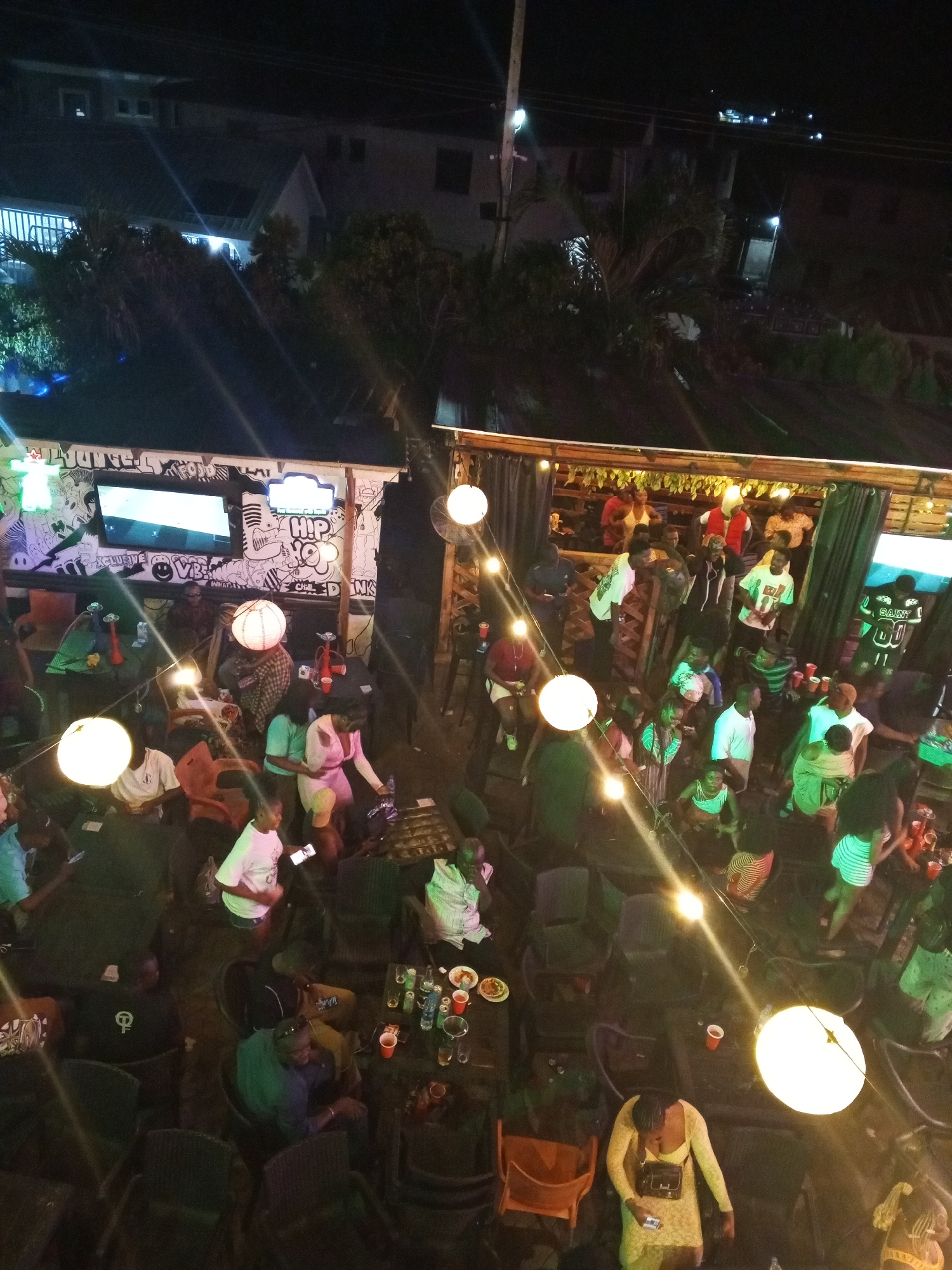 The image shows a nighttime view of an outdoor bar or club setting in an urban environment. There are many people present, mostly adults, some standing, some seated at tables. They appear to be socializing and consuming food and drinks. There are hanging string lights with round bulbs illuminating the scene. In the background, there are buildings, and on the left side, there is a structure with a TV screen and graffiti-style art on the wall.