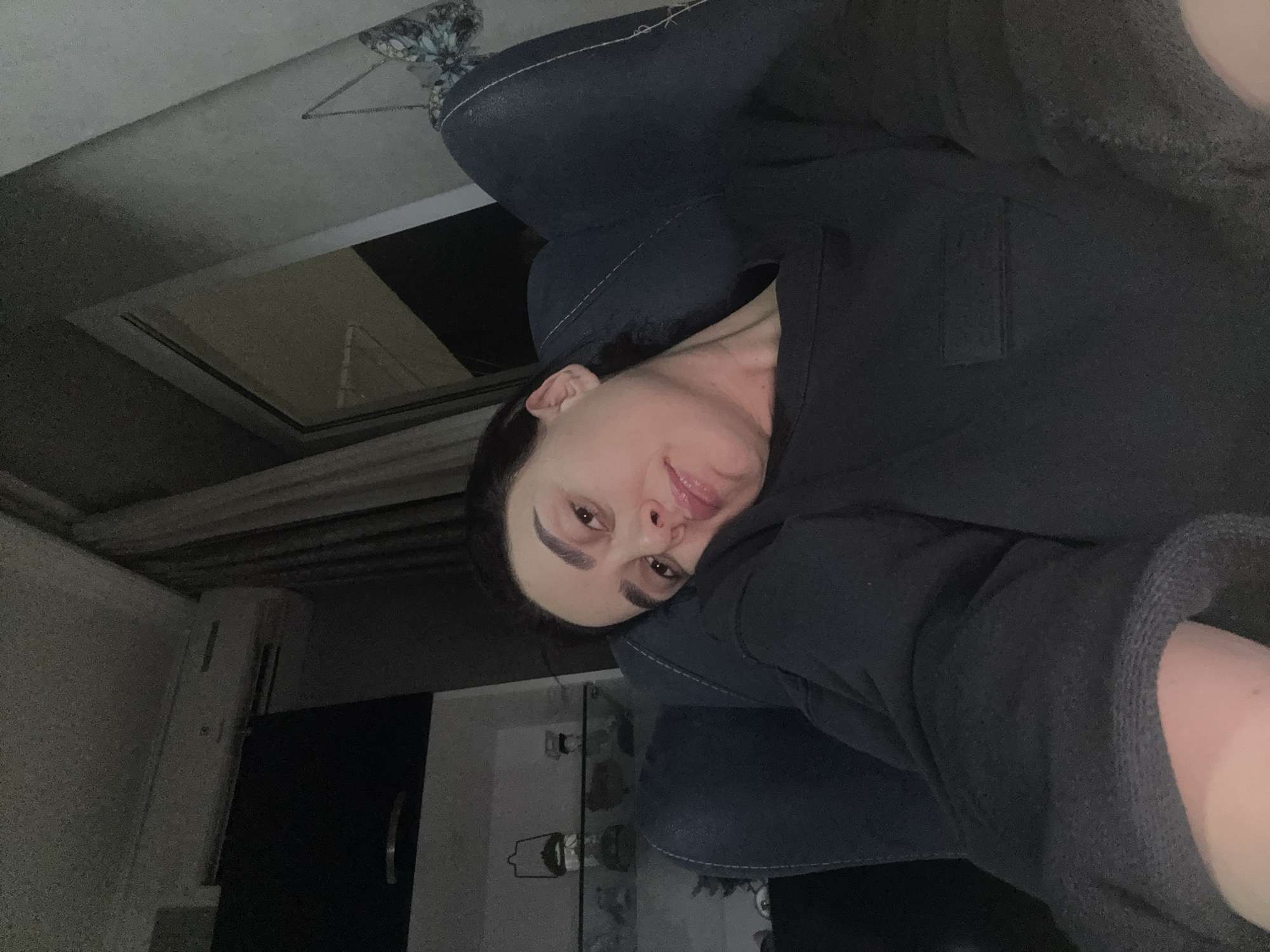 This indoor image features a woman posing for a selfie in a dimly lit room, likely at night or in the evening. The main subject is a woman with dark hair, well-defined eyebrows, and light skin, looking directly at the camera with a slight smile. She is wearing a dark, possibly black or charcoal grey, long-sleeved top or hoodie, which covers most of her body, with a part of her arm visible on the right side of the frame.
She appears to be leaning against or sitting in a piece of furniture, suggested by a dark blue or black upholstered surface with white stitching visible behind her head and shoulder.
The background reveals parts of a room. To the left of the woman (viewer's left, if the image were upright), there's a reflection of what might be a window or light source, and a patterned fabric (possibly floral) on an unseen object, perhaps a pillow or part of the furniture. To her right (viewer's right, if upright), a white air conditioning unit is mounted on a light-colored wall, next to a partially visible dark door with a silver handle. Above this area, some wooden beams or ceiling structure can be seen. The low lighting emphasizes the woman's face while keeping the rest of the room in shadow. No visible text is present. The scene suggests a relaxed, personal moment in a home or private accommodation in Tbilisi, Georgia.
