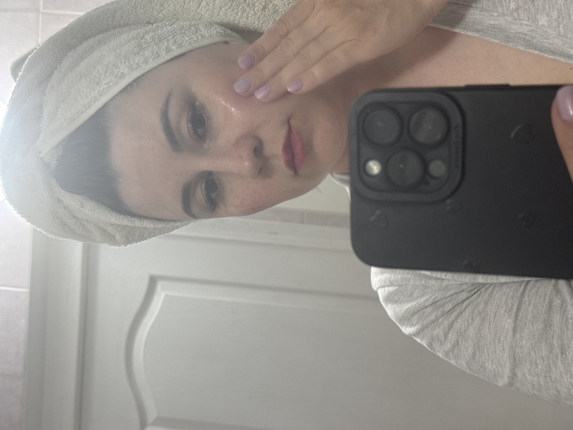 A person wearing a towel on their head and a light grey shirt is taking a selfie. The person's hand is near their face as if applying lotion. The background shows a white door with a patterned design. The location is not possible to determine from the image, as there are no recognizable landmarks or geographical information to determine Zav'yalovo, Russia.