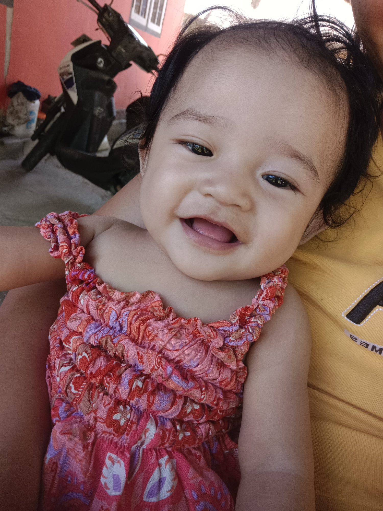 An overhead close-up photograph shows a smiling baby in a pink and red floral print dress. The baby's head is turned slightly towards the viewer, with dark hair and dark eyes. The baby's mouth is open, revealing a pink tongue. The baby is being held by someone wearing a yellow shirt with black lettering on the right side of the frame. In the blurred background, a red wall and a motorcycle are visible, suggesting an outdoor setting, potentially a residential area in Kawua, Indonesia. The ground appears to be concrete.
