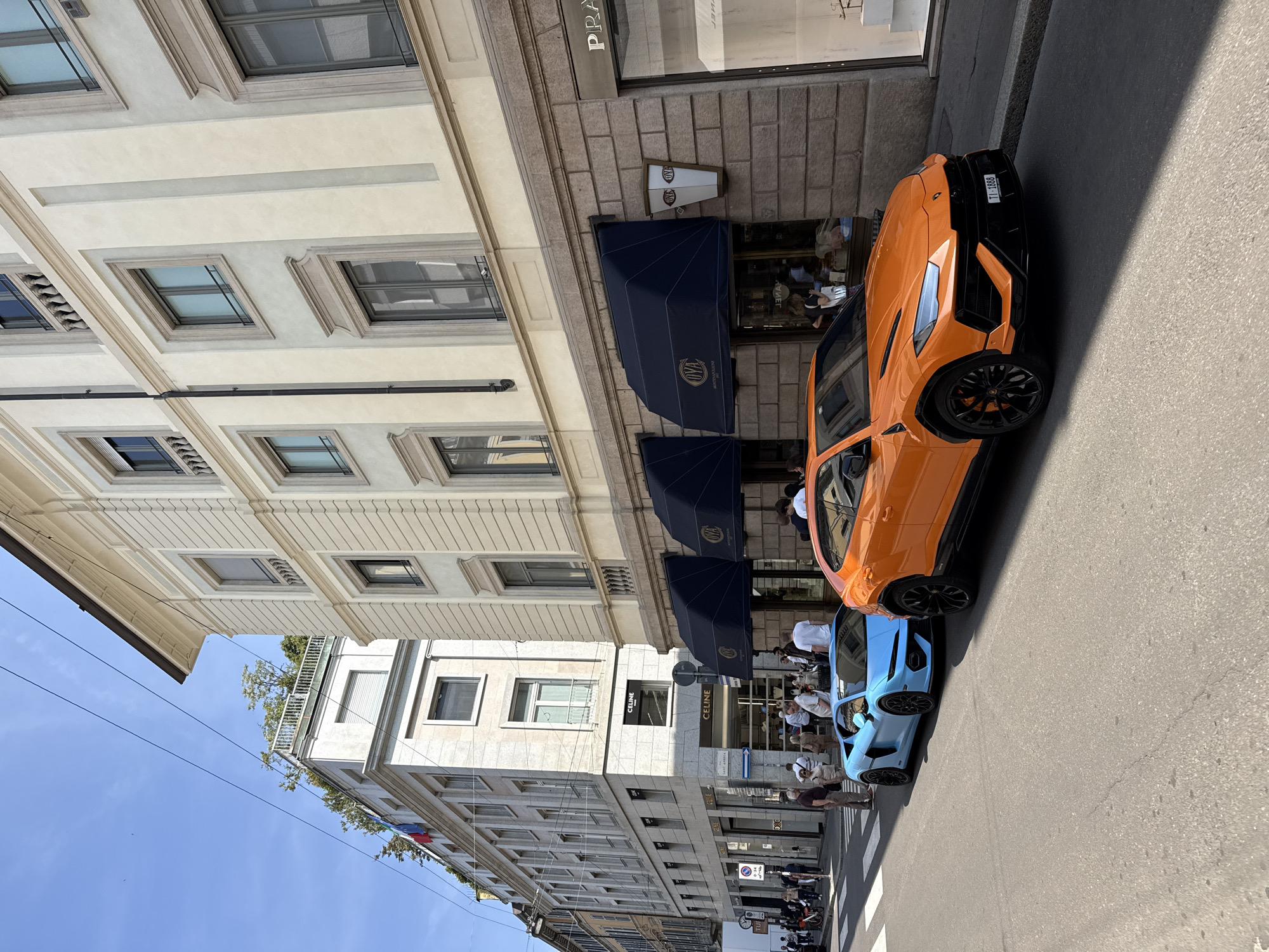 An eye-level outdoor shot depicts a street scene in Milan, Italy, featuring parked luxury vehicles and storefronts. An orange Lamborghini Urus is parked at an angle, followed by a light blue vehicle, possibly a smaller sports car, also parked at an angle. Several individuals are visible on the sidewalk, some standing near the vehicles and others walking along the street. Two visible businesses are CELINE and PRADA, recognizable by their signage and storefront awnings. The architecture consists of multi-story buildings with traditional windows. The street appears to be in a high-end retail area, with well-maintained sidewalks. The sky is blue, indicating daytime. Shadows are cast on the road and buildings, suggesting bright sunlight.